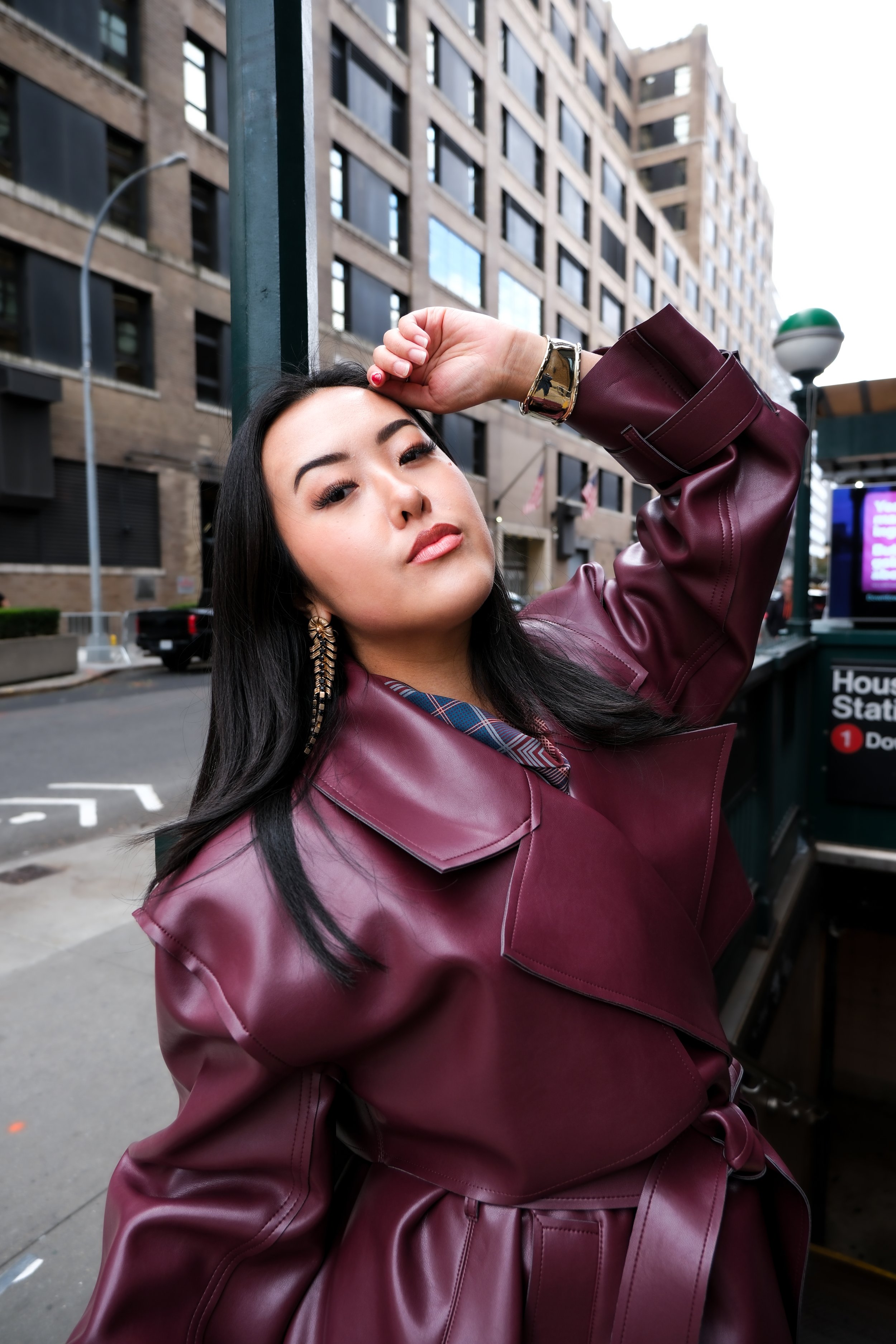 A woman in a maroon leather coat standing outdoors on an urban street, posing with her arm raised and hand on her forehead, with tall buildings and a subway entrance in the background.