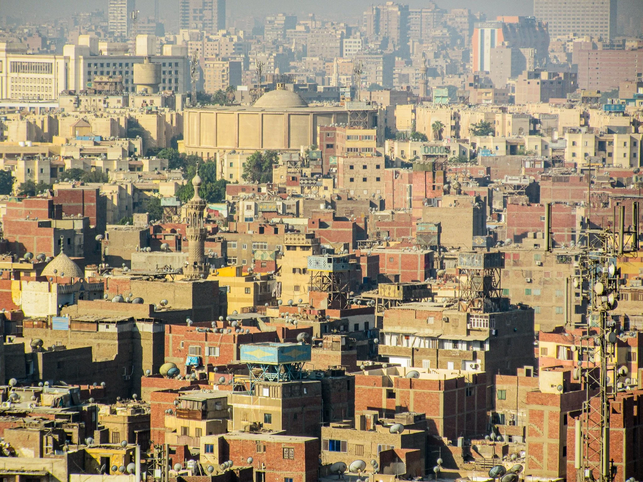 Cityscape of a densely packed urban area with a variety of buildings, rooftops with antennas and satellite dishes, and a large domed building in the distance, under a hazy sky.