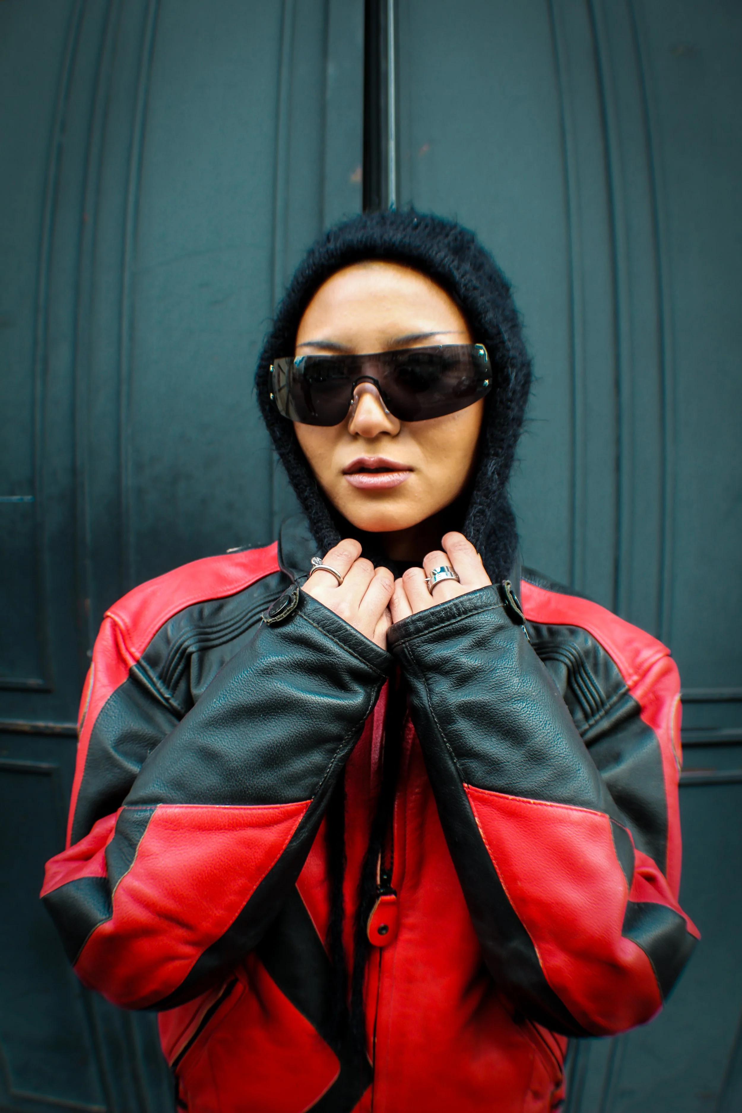 A woman wearing a black hoodie, oversized sunglasses, a red and black leather jacket, and rings, standing in front of a dark green door.