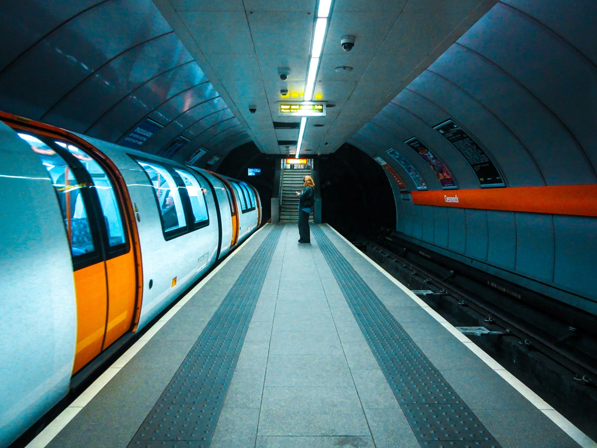 An underground metro station with a woman standing on the platform looking at her phone, train arriving on the left, station name 'Crossrod' on the side wall, stairs at the far end.