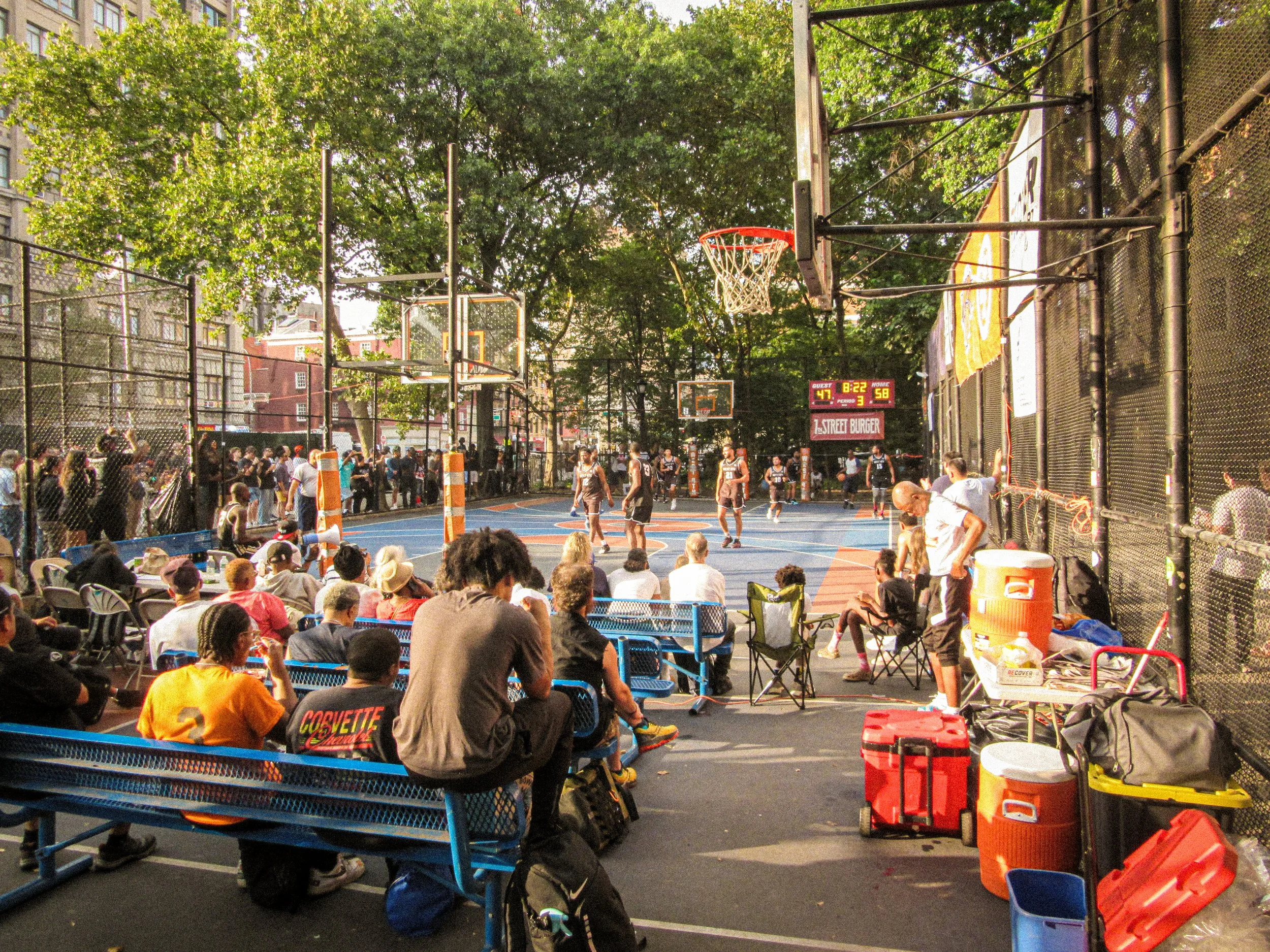 People watching a basketball game on an outdoor court surrounded by tall fence panels and trees in a park.