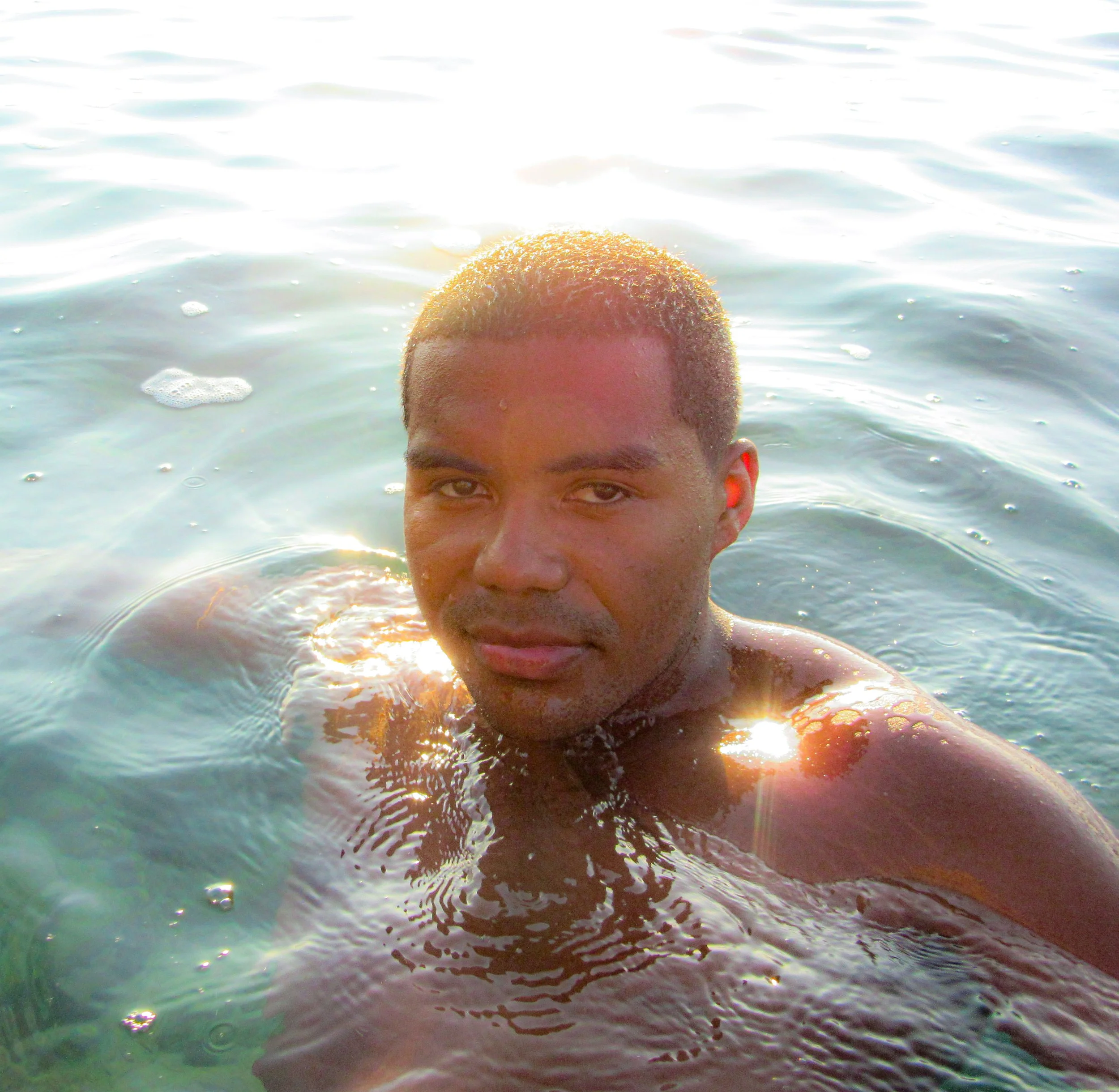 A man with short, textured hair swimming in a body of water during sunset, with sunlight reflecting on the water and on his face.
