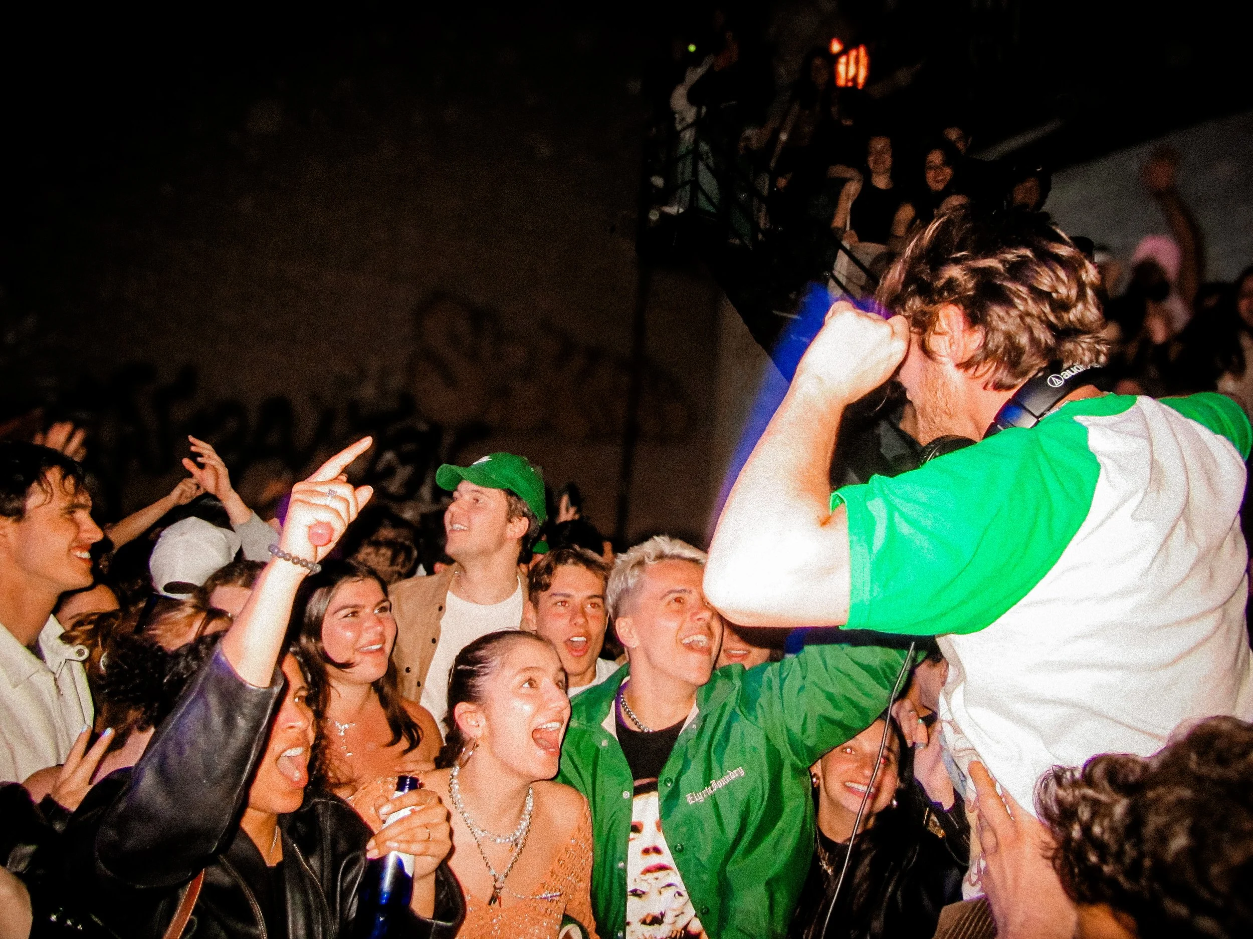 A DJ in a green and white shirt, wearing headphones, appears to be performing at a lively party or concert. The crowd around him is excited, smiling, and raising their hands, enjoying the event.