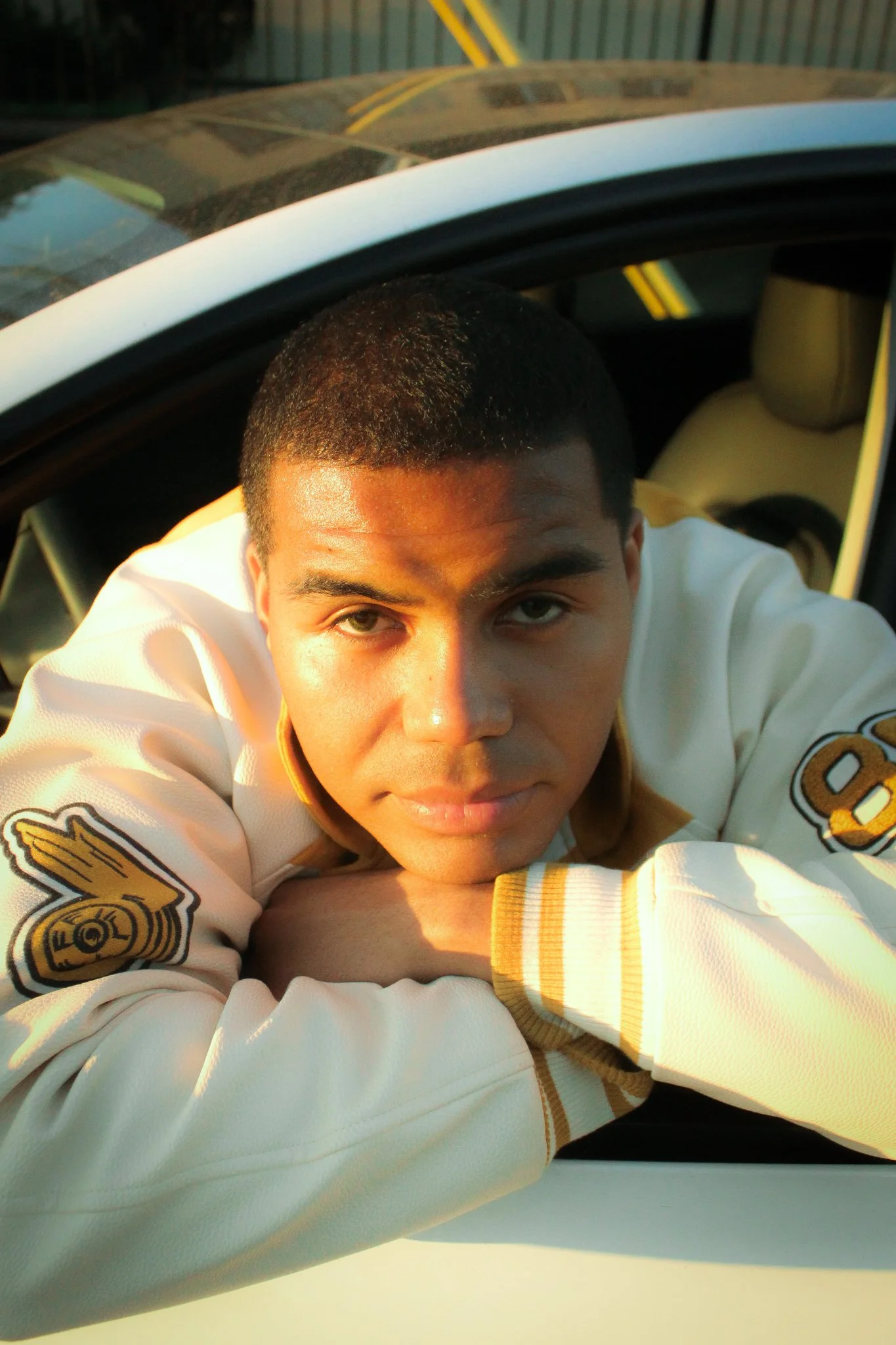 A young man with short black hair leaning out of a car window, wearing a white and gold varsity jacket, with a serious expression.