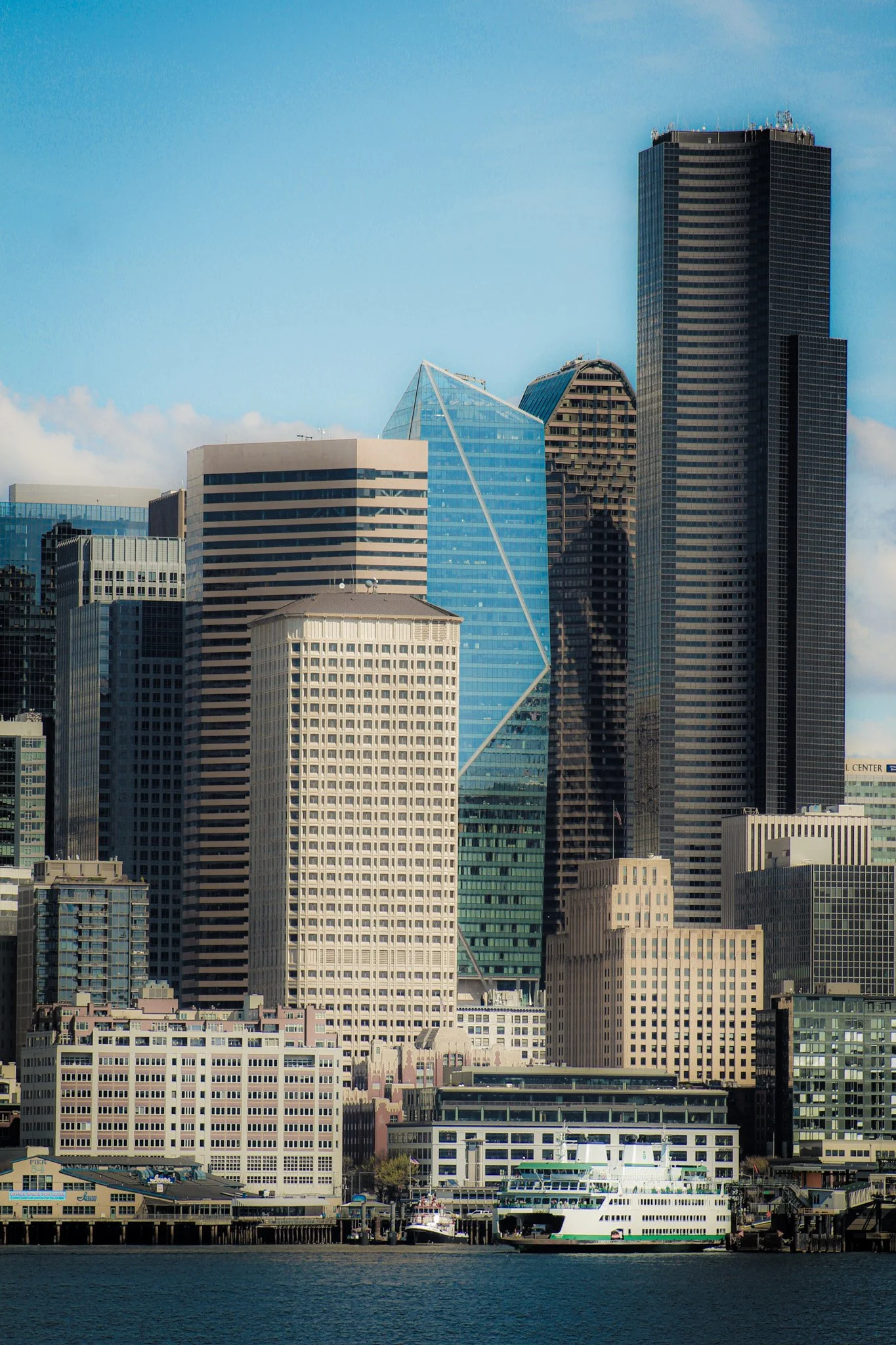 A city skyline featuring tall skyscrapers, including a blue glass building with a diagonal white line, under a blue sky with some clouds, and a body of water with boats in the foreground.