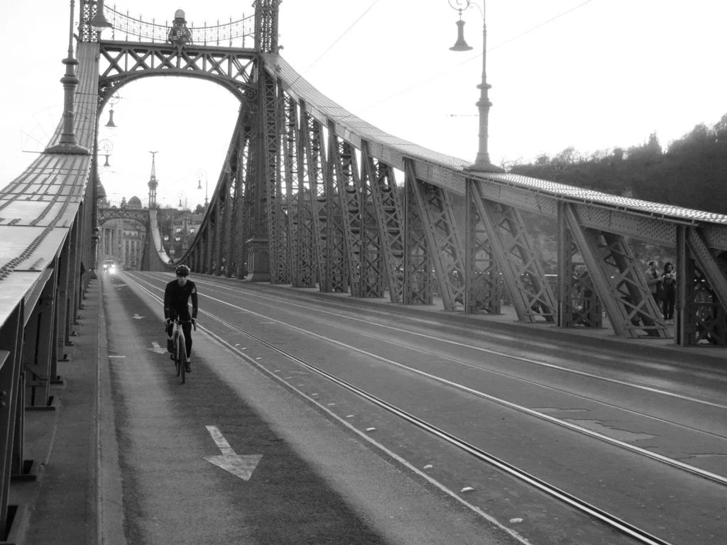 A cyclist riding on a bike lane on a bridge with tram tracks, with historic lamp posts and a large suspension bridge structure in the background, in black and white.