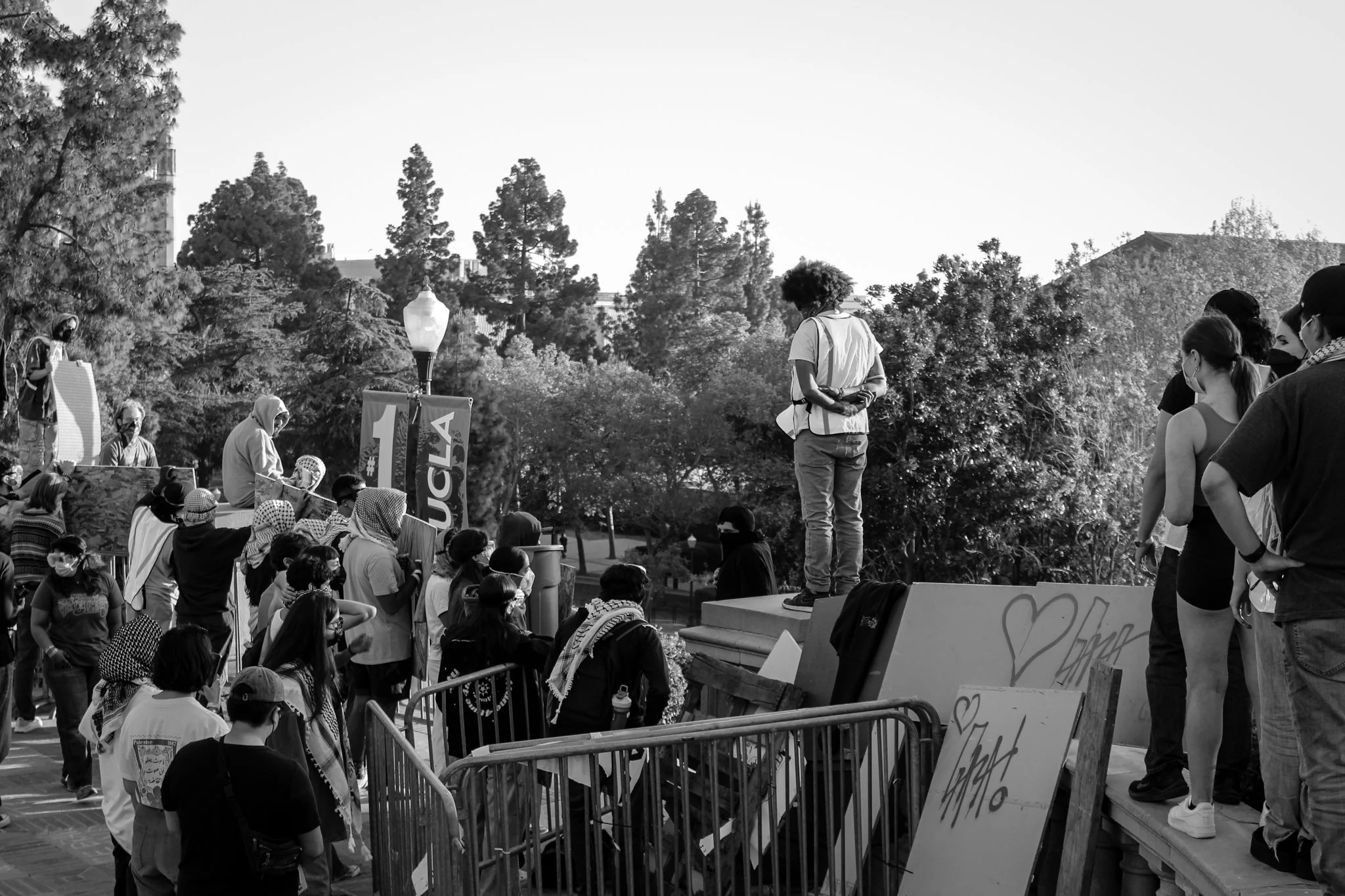 A black and white photo capturing a public gathering or protest outdoors, with a crowd of people, some wearing masks. There are speakers speaking on a stage, with protest signs and banners visible, including one that says 'BLACK'. Trees and buildings