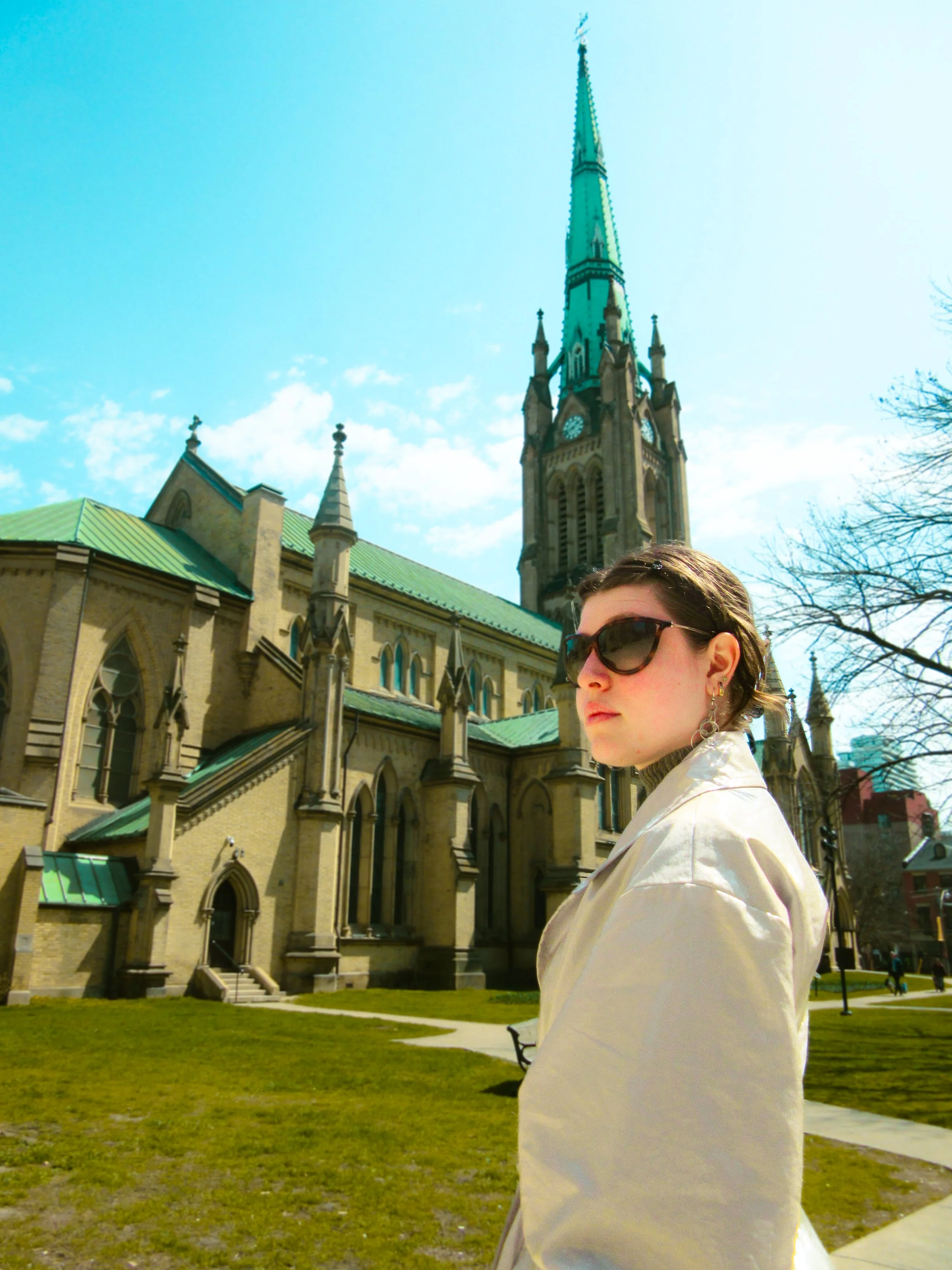 Woman in sunglasses standing outside a historic church with green roof and steeple under blue sky.