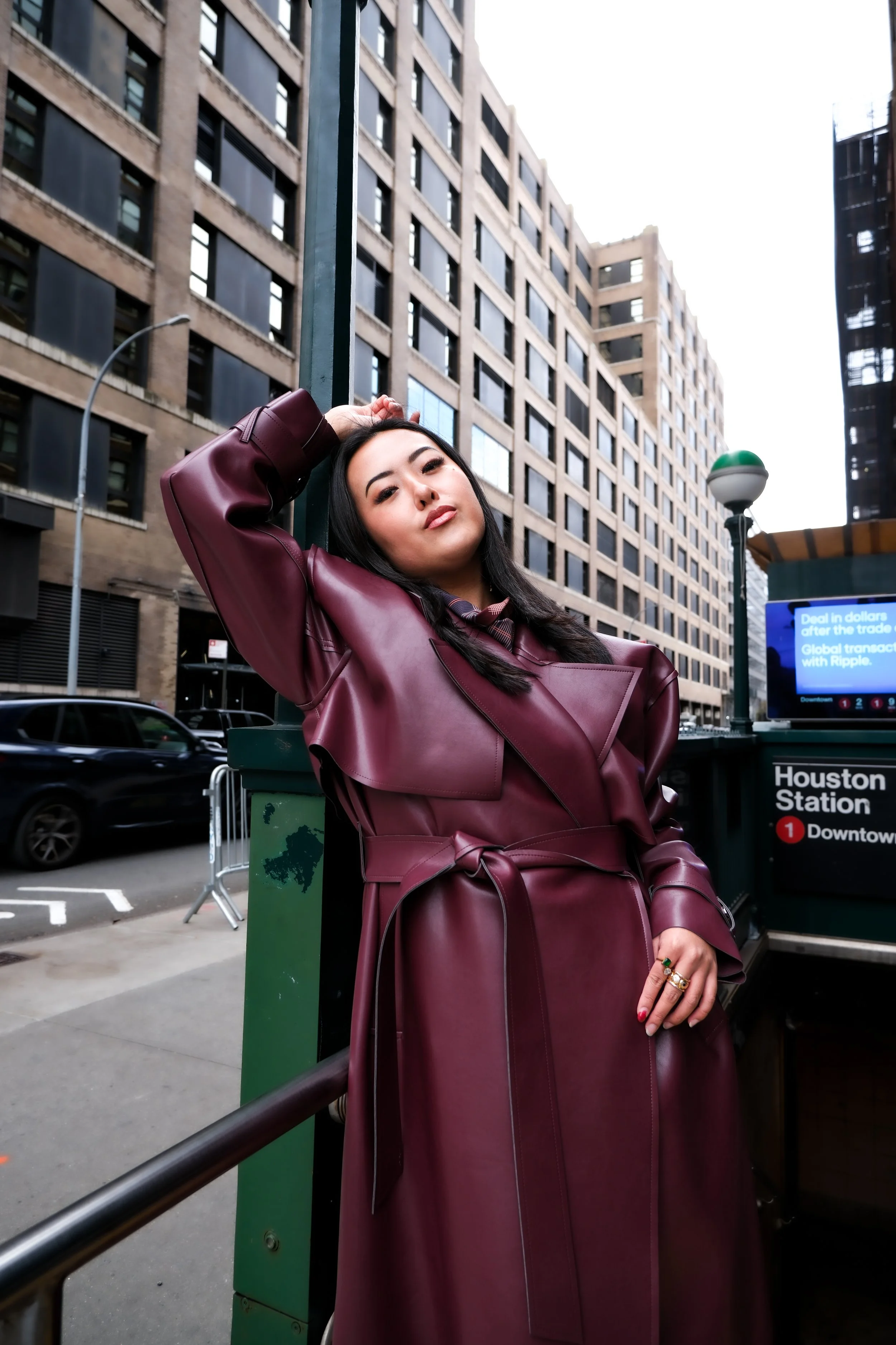 A woman in a maroon leather trench coat leaning against a green pole outside an urban subway station with tall buildings in the background.