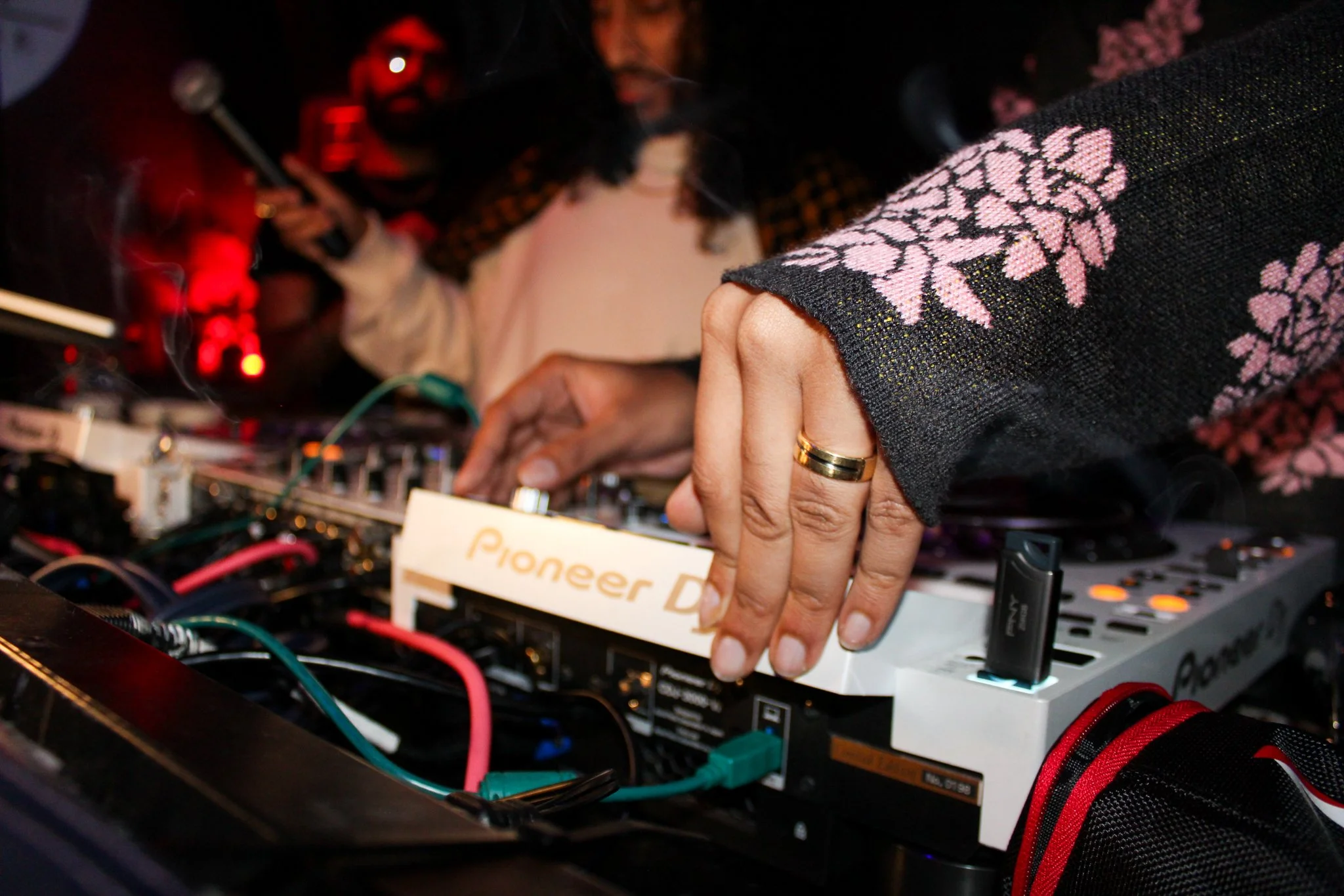 Close-up of a DJ's hand with a gold ring adjusting a music mixer. The DJ is wearing a black jacket with pink floral patterns on the sleeve. In the background, there are two people, one holding a microphone, with red and black lighting creating a live
