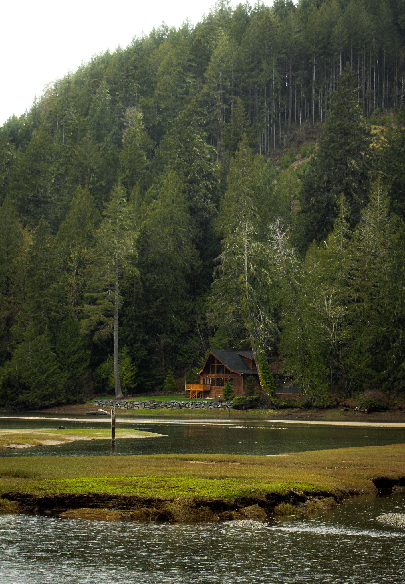 A wooden cabin near a body of water, surrounded by dense green trees and hills.