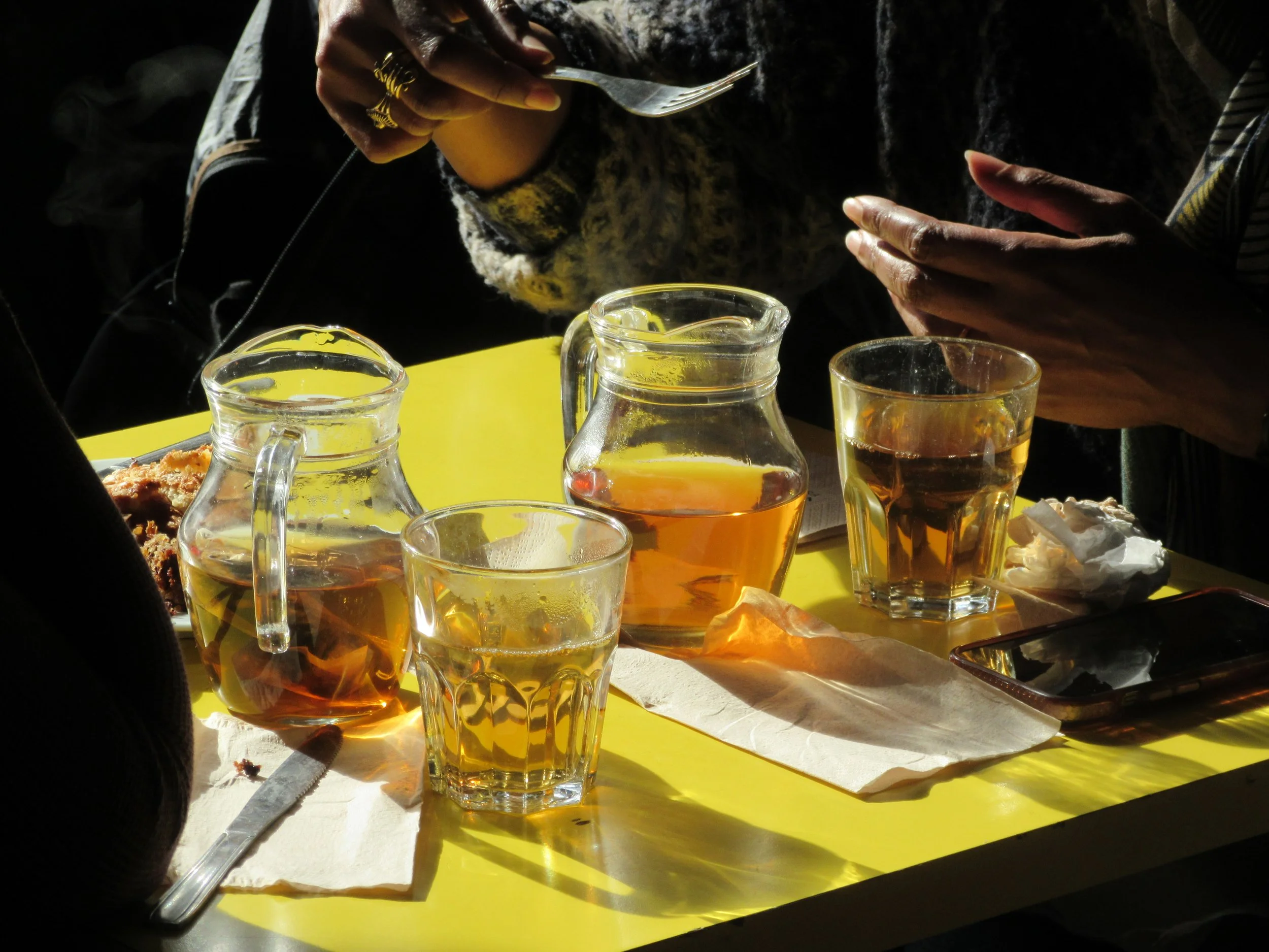 A table with three glass pitchers and three glasses filled with tea, along with paper napkins, a spoon, and a plate of fried food, with people sitting around it in sunlight.