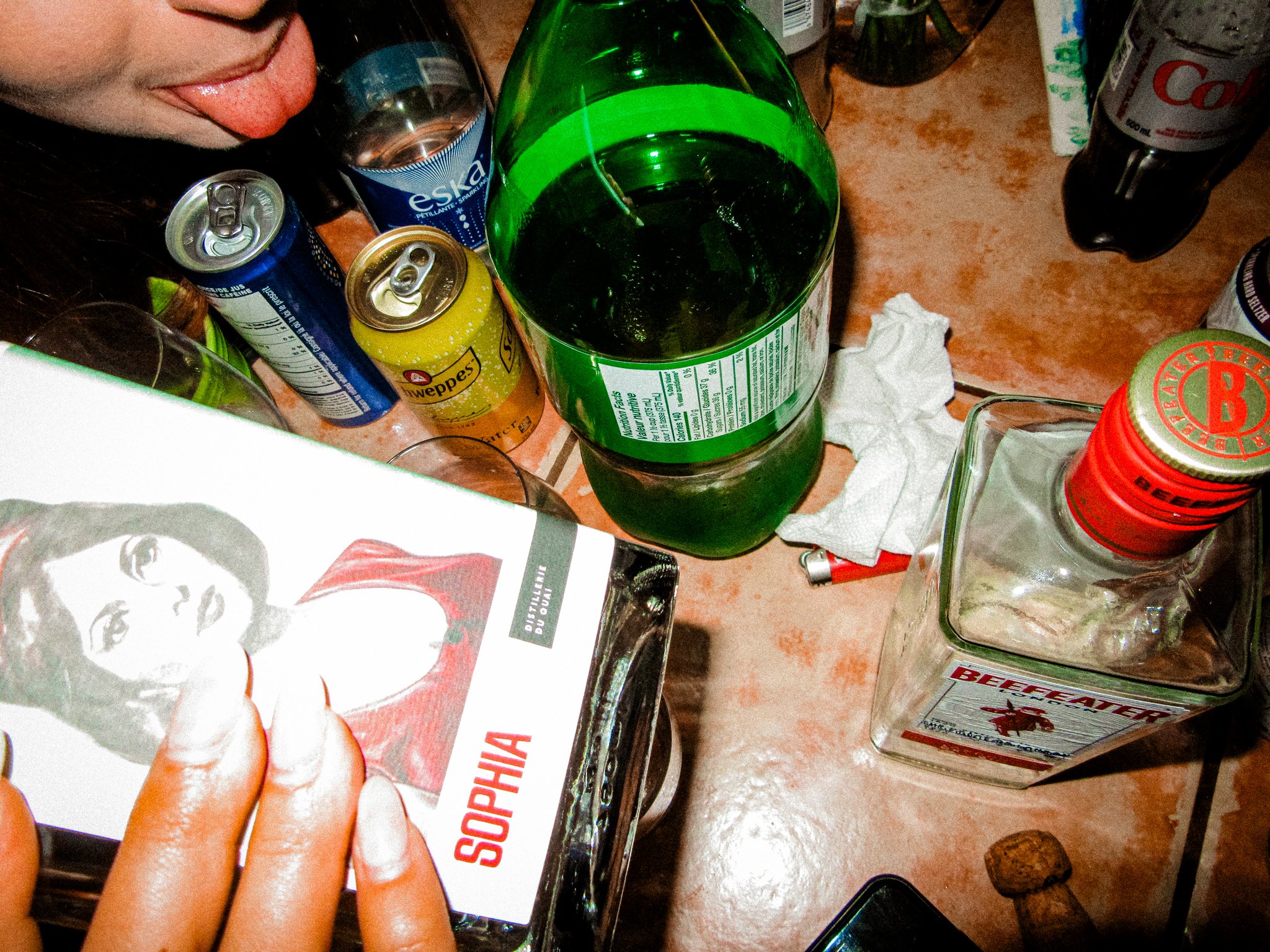 A cluttered table with various unopened soda cans, a partially consumed green soda bottle, a bottle of Beefeater gin, a pack of Sophia cigarettes, and a woman sticking out her tongue in the top left corner.