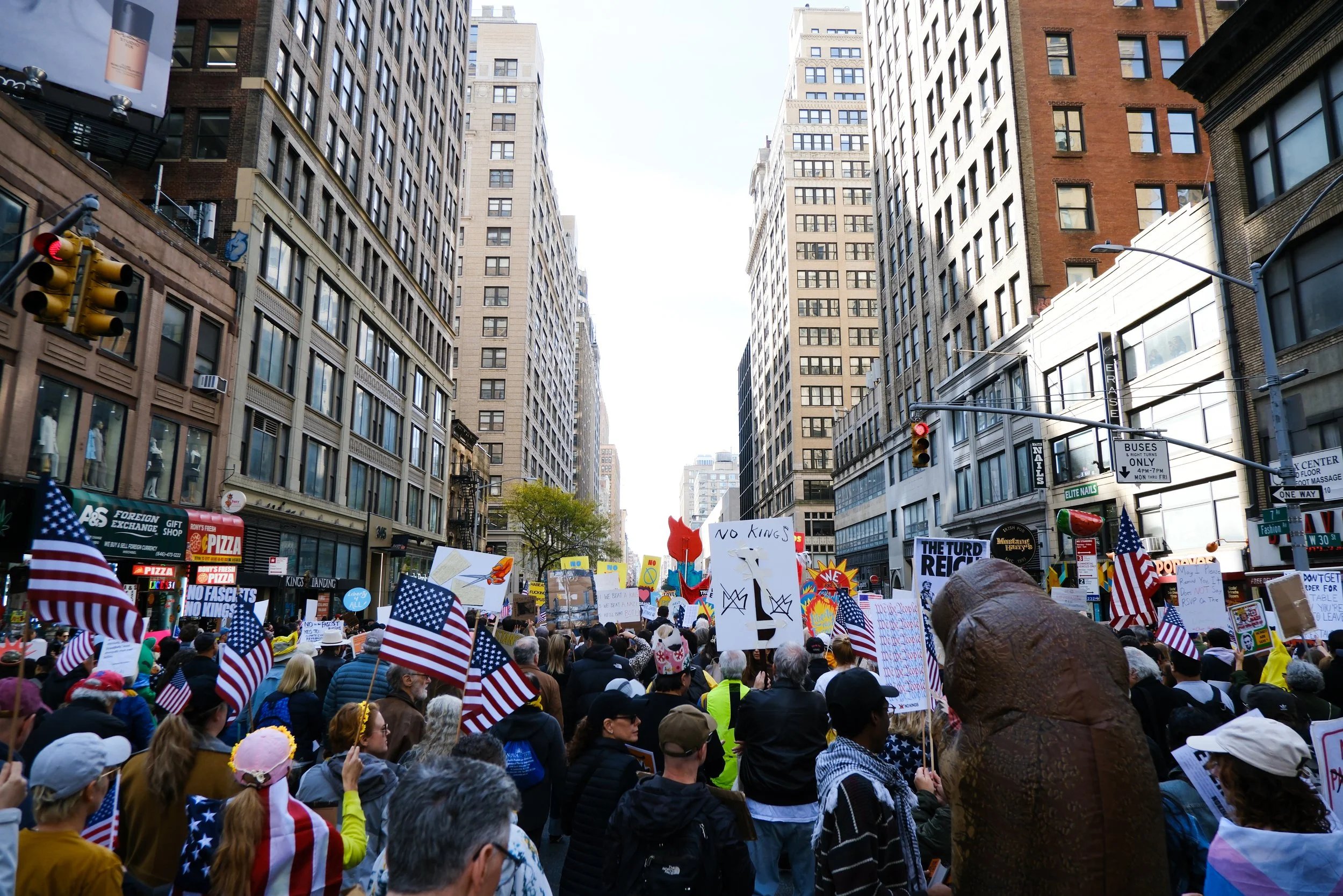 Crowd of people protesting on a city street, holding various signs and American flags, surrounded by tall buildings.