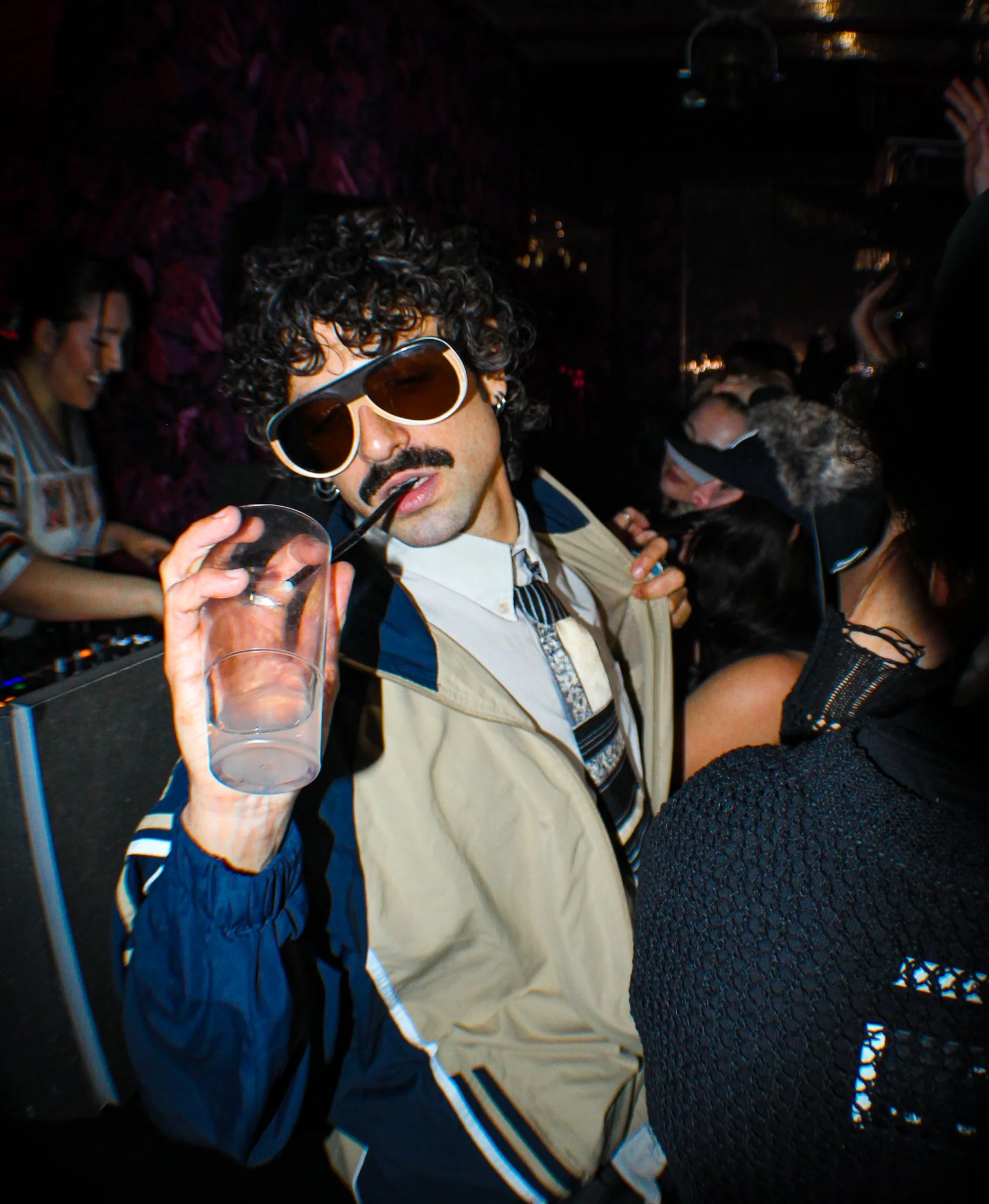 Man with curly hair, sunglasses, and mustache in a party environment, holding a glass with a straw, surrounded by people in a lively setting.