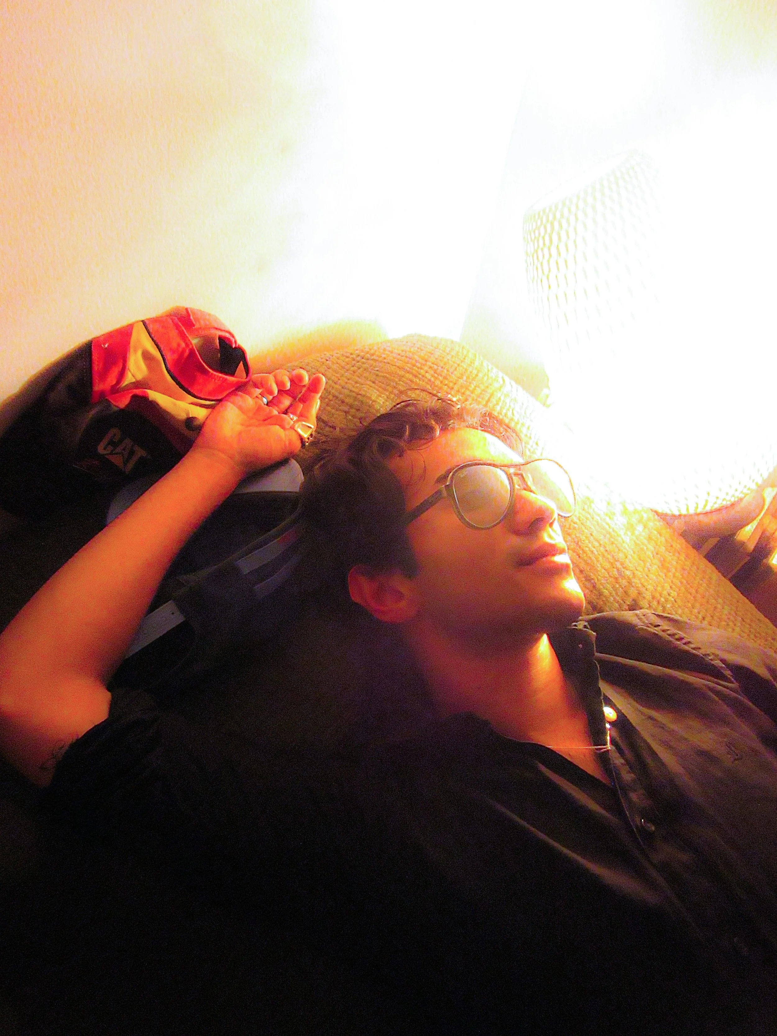A person with glasses lying on a couch, face illuminated by a bright lamp, with a pillow behind their head and a backpack nearby.