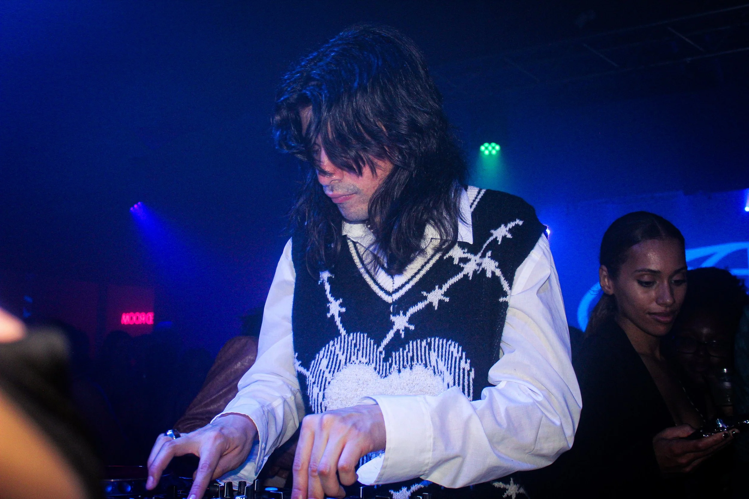 A man with long dark hair mixing music at a DJ booth in a club, wearing a sweater vest over a white shirt, with people and colorful lights in the background.