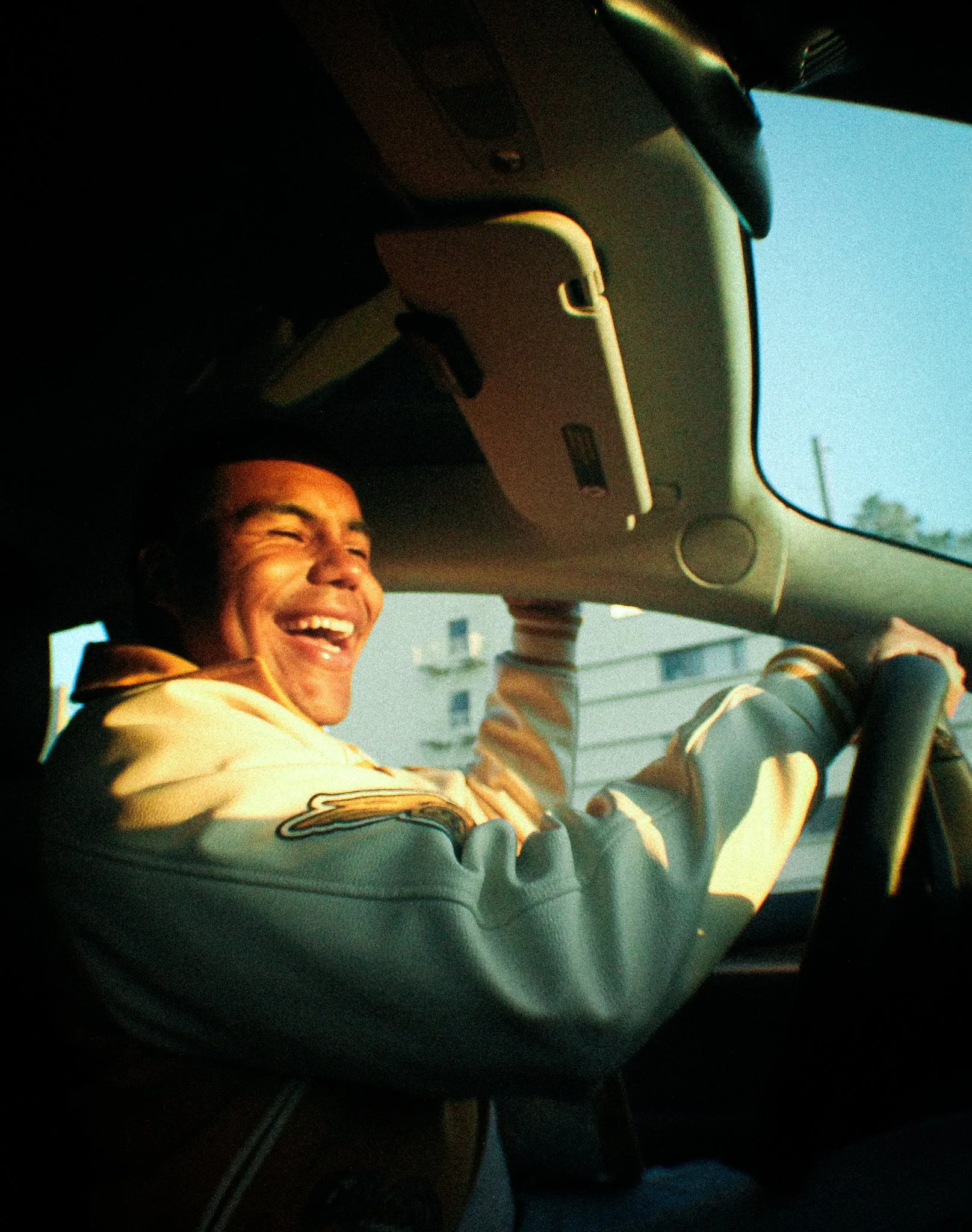 Person smiling and enjoying the drive in a vehicle with sunlight on face, wearing a jacket with a patch on the sleeve.