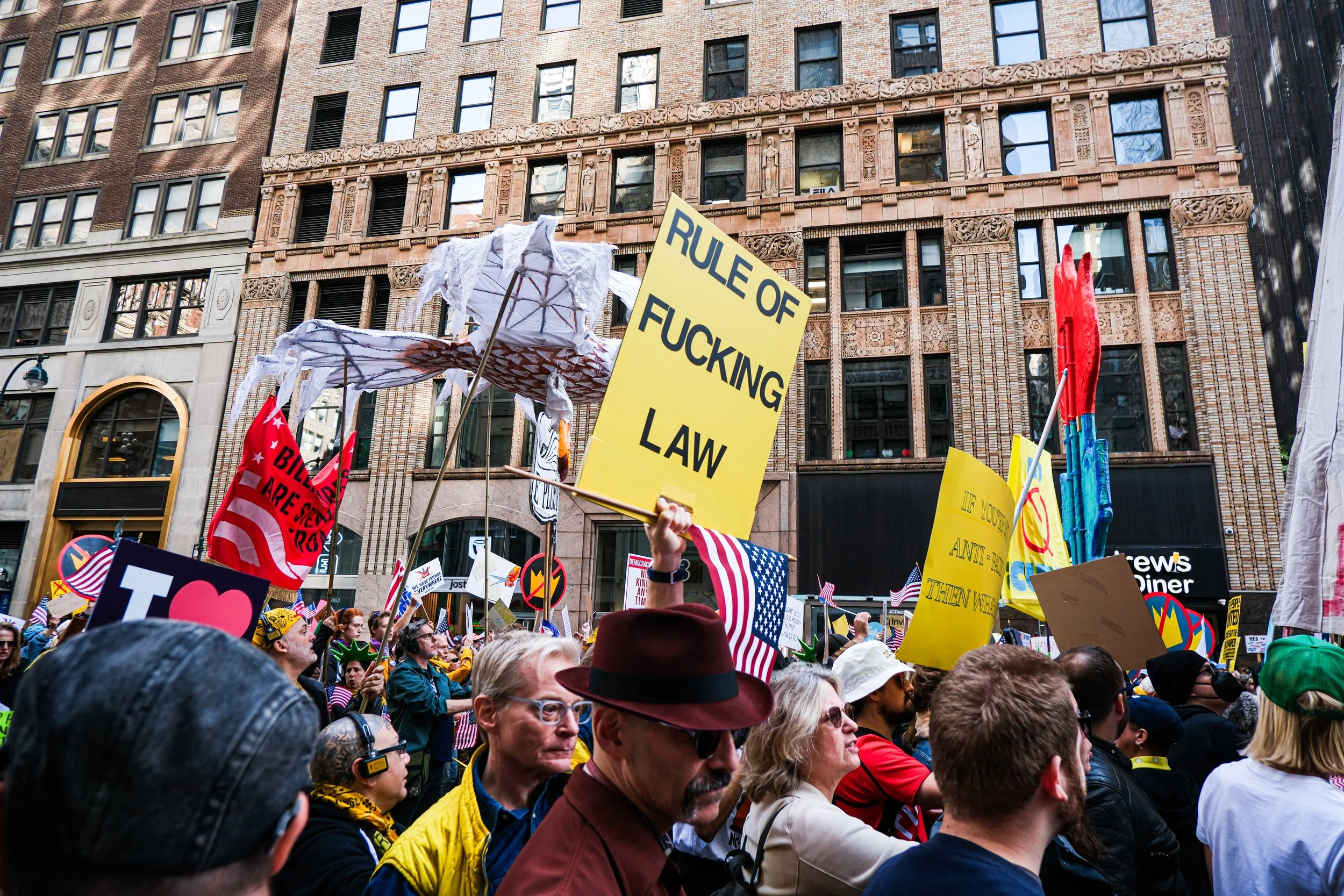Protest crowd in an urban area holding signs, including one that says 'Rule of F***ing Law,' with diverse participants and historic buildings in the background.