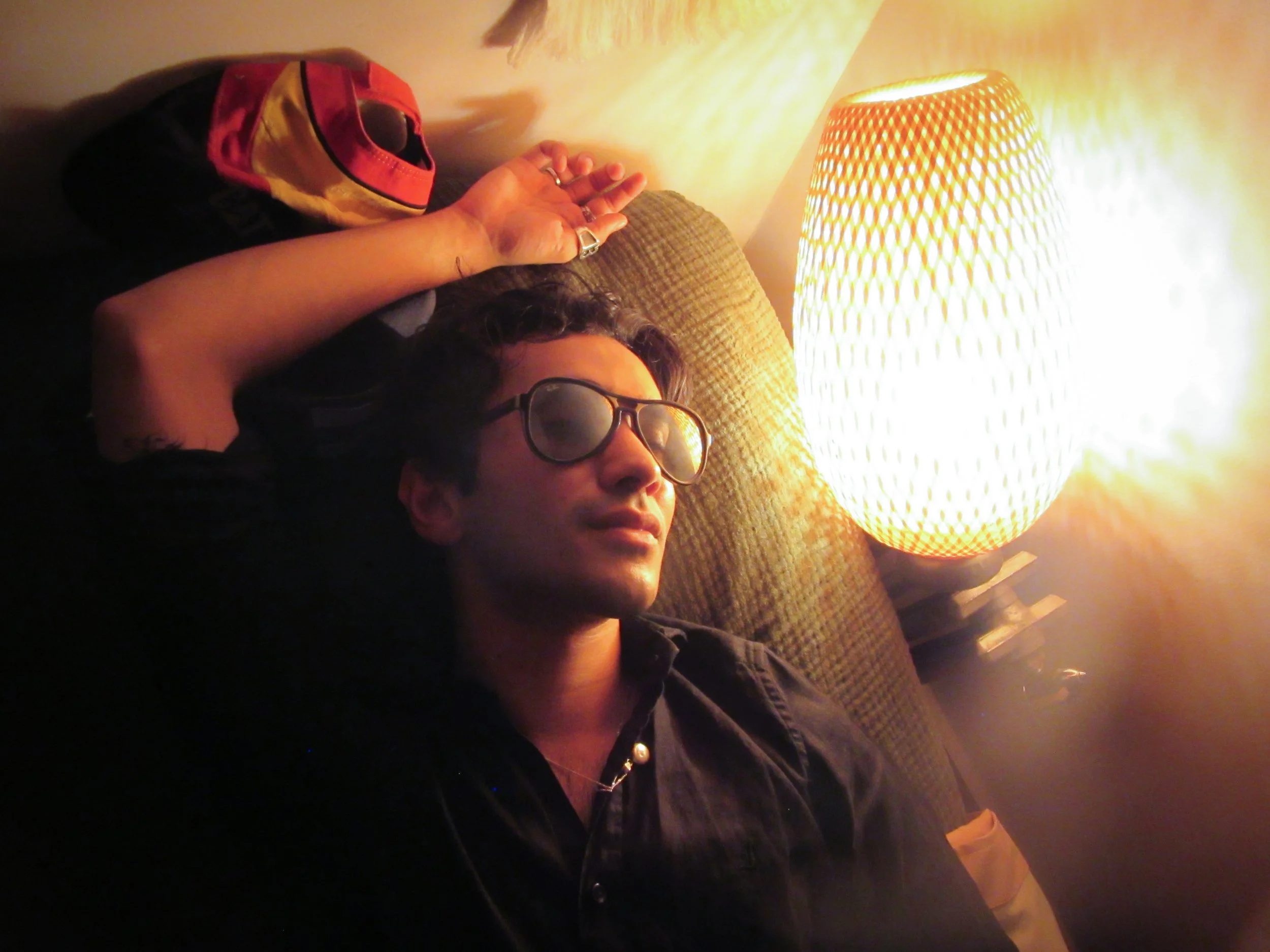 A person with dark hair, wearing glasses, relaxing with eyes closed on a couch next to a lit lamp, with a colorful hat resting behind their head.