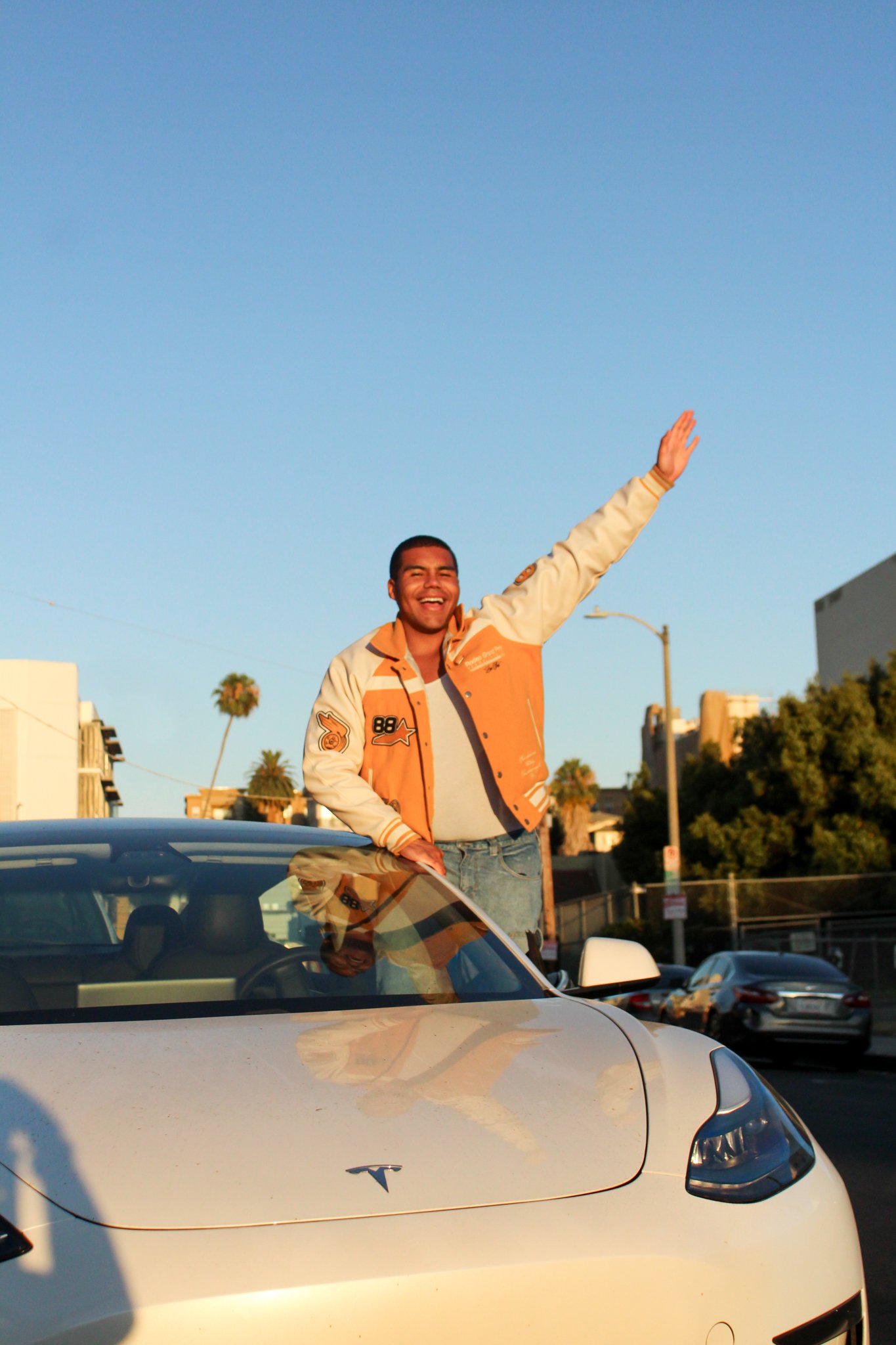 A man standing in the open sunroof of a white Tesla car, smiling and waving, during sunset with a clear blue sky in the background.
