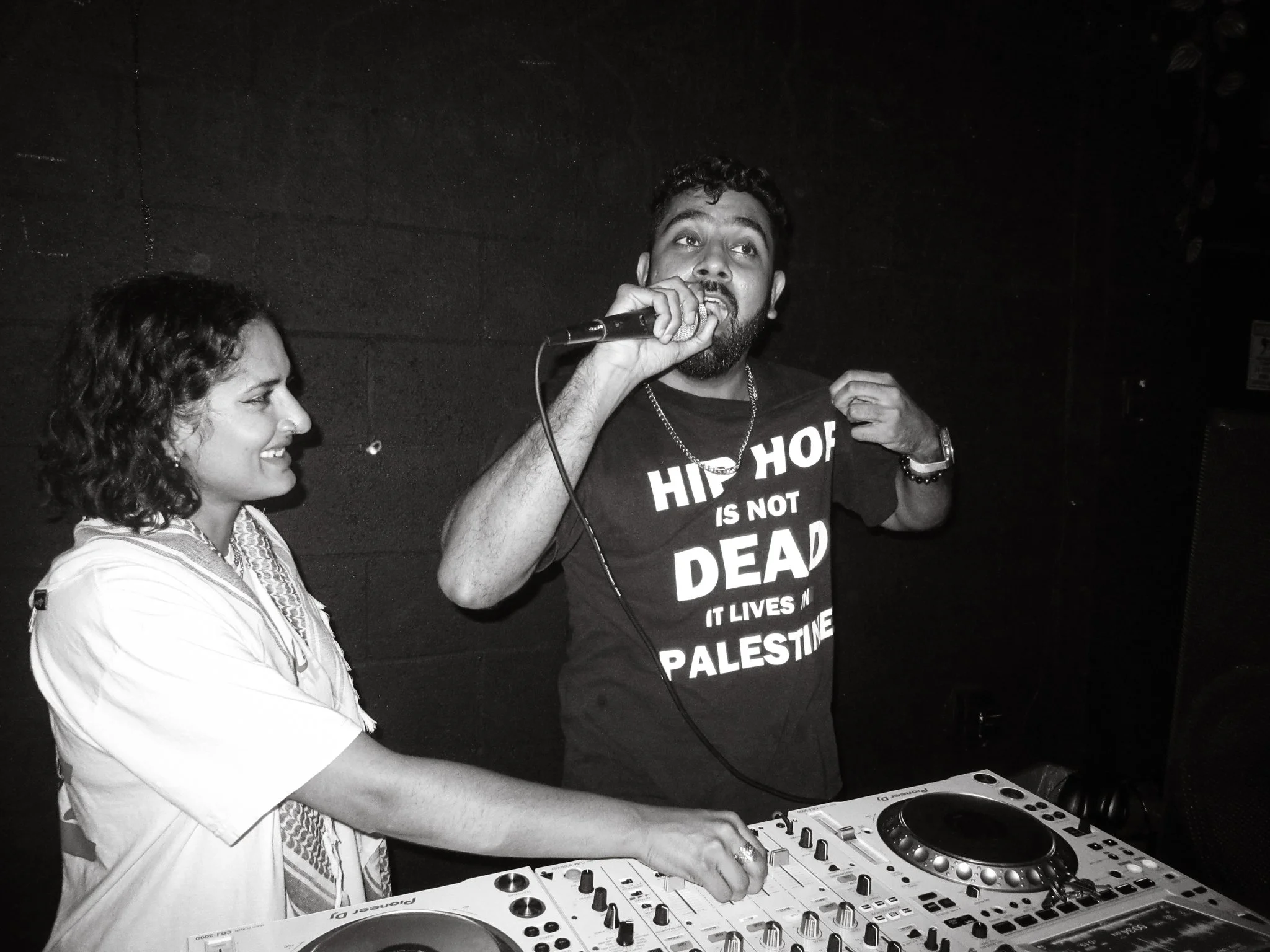 A woman and a man are DJing; the woman is smiling at the controls, while the man is speaking into a microphone, wearing a T-shirt that says 'Hiphop is not dead, it lives in Palestine'.