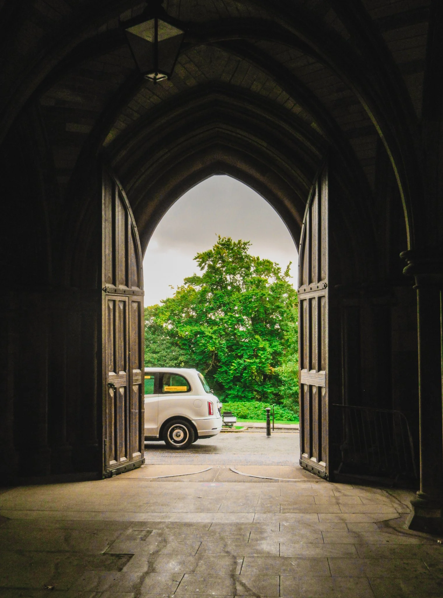Open wooden church doors leading to a parking lot with a white Rolls Royce and green trees in the background.