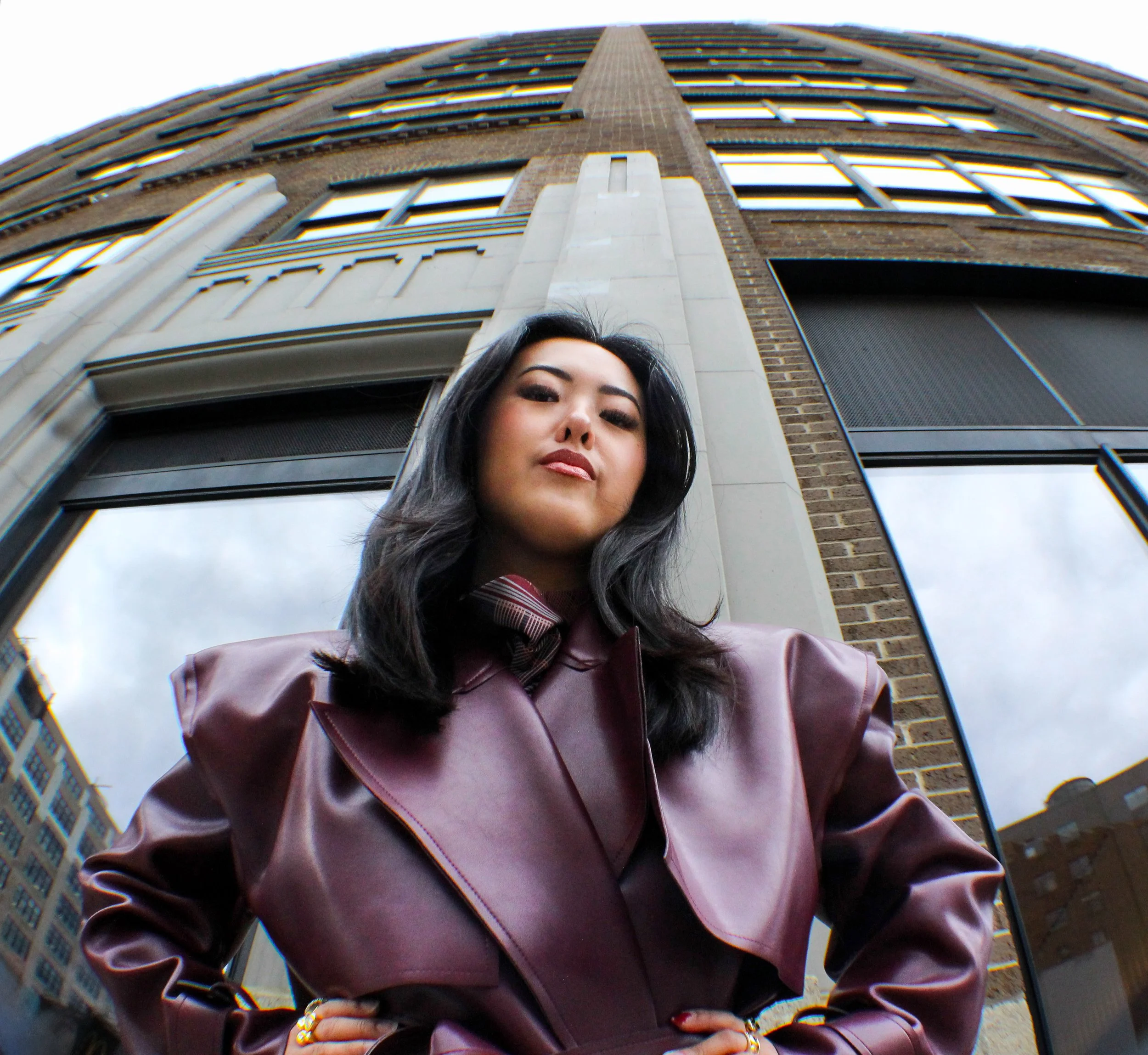 A confident woman in a leather jacket standing in front of a tall building, captured from a low-angle with a fisheye lens.