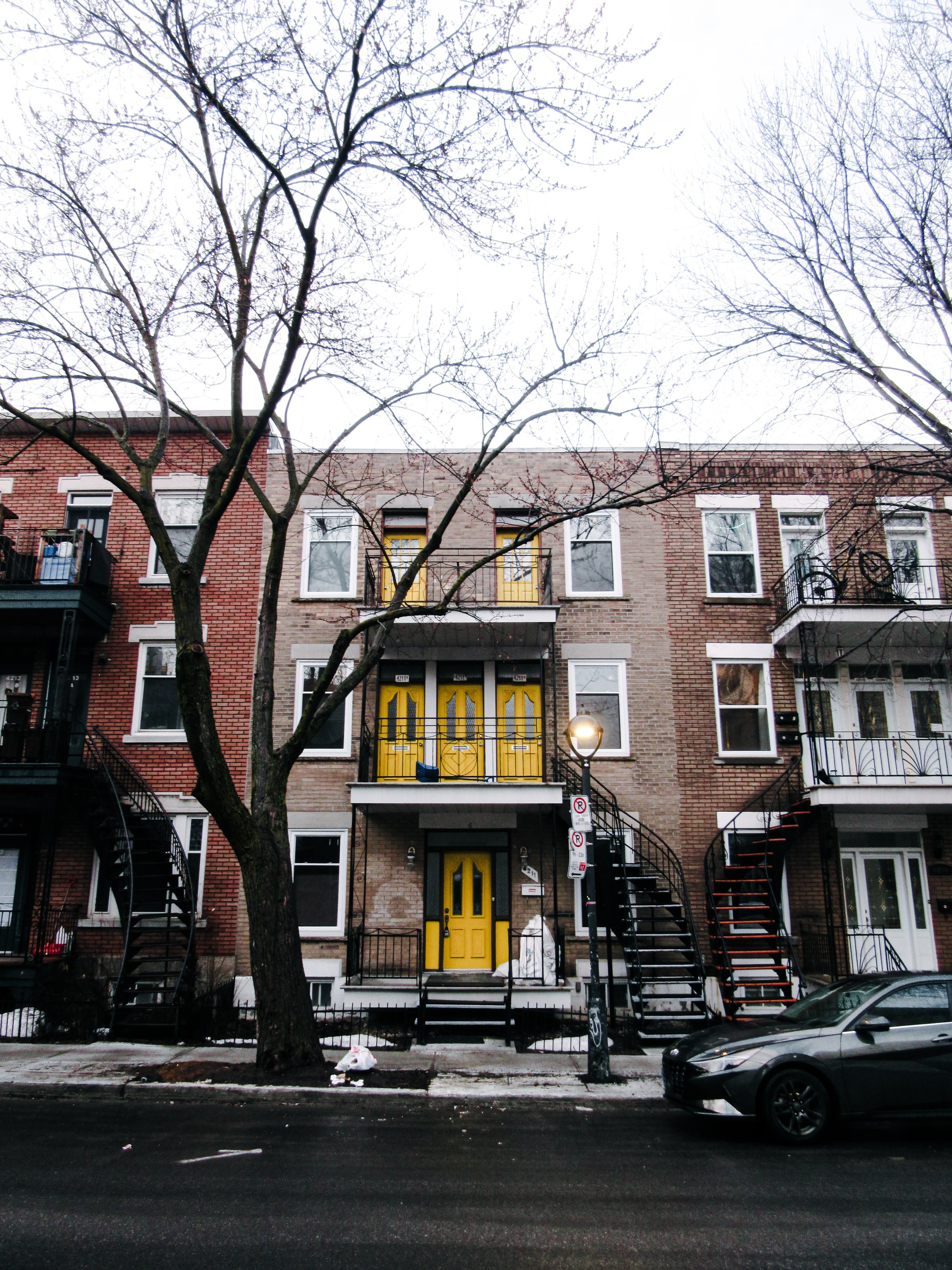 A residential building with three floors, brick exterior, and yellow doors. There are stairs on both sides leading to upper balconies. A large tree is in front, and a car is parked on the street.
