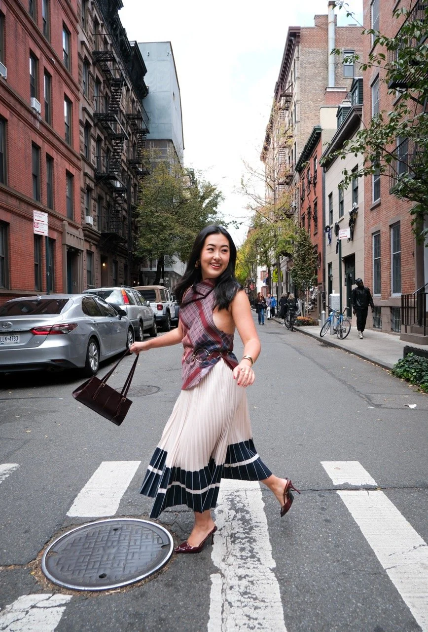 A woman crossing a city street at a crosswalk, smiling, carrying a handbag, dressed in a pleated skirt, high heels, and a sleeveless top, with cars parked along the street and buildings lining the background.