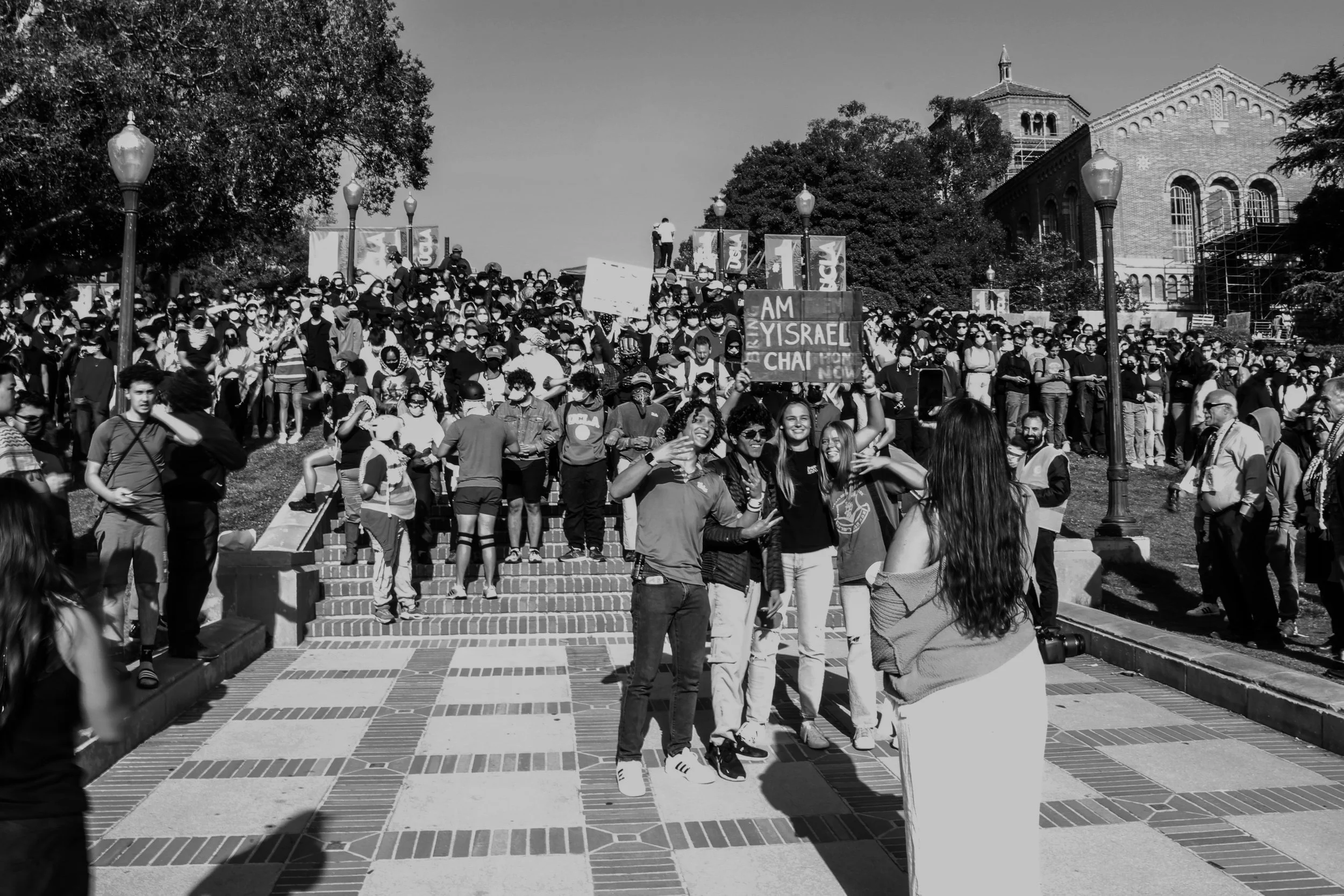 A large crowd of people gathered outdoors on a sunny day, with some holding signs. The crowd appears to be participating in a rally or demonstration. The people are standing on steps and the surrounding area has trees, lampposts, and historic buildin