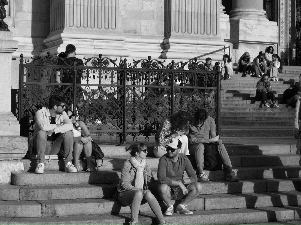 People sitting on steps outside a historic building with ornate columns and railing, some engaged in conversation, reading, or looking at phones.
