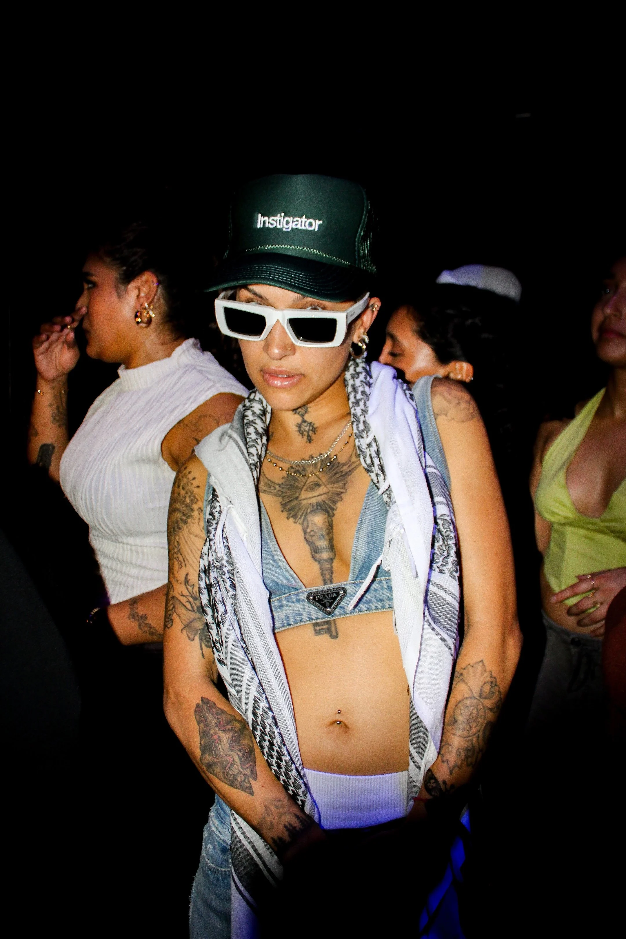 A woman with tattoos and piercings wearing a black cap labeled 'Instigator', white sunglasses, layered necklaces, a denim bra, and a sleeveless shirt, at a nightclub or party with other women in the background.