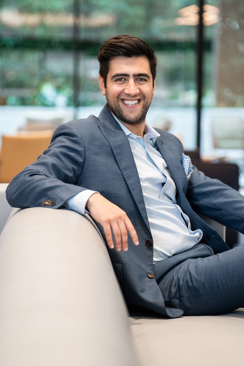 Smiling man in a gray suit sitting on a white couch in a modern office or lounge area.