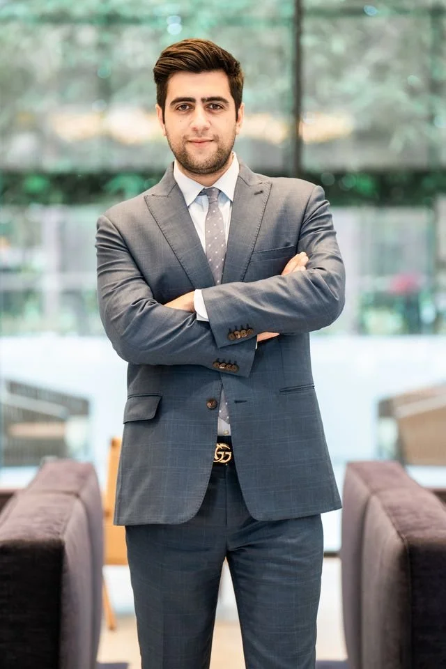 A young man in a gray suit standing with arms crossed in an indoor setting with large windows and blurred couches in the background.