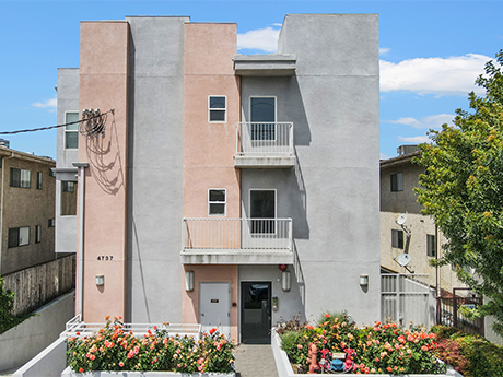 Colorful three-story apartment building with balconies, surrounded by flowers and trees, under a partly cloudy sky.
