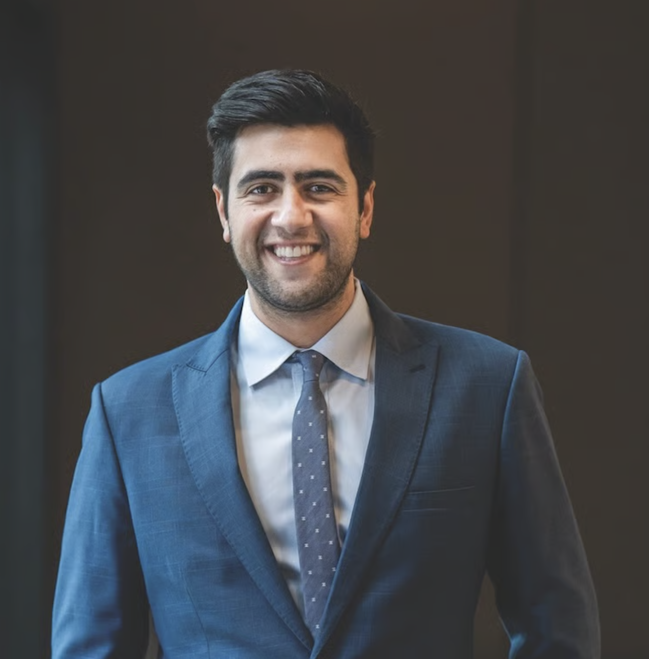 A young man in a dark blue business suit, white shirt, and navy tie, smiling and standing against a dark background.