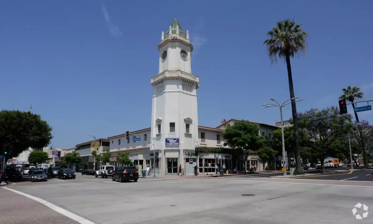 A white clock tower with a green-tiled spire at a busy intersection, surrounded by palm trees and buildings on a sunny day.