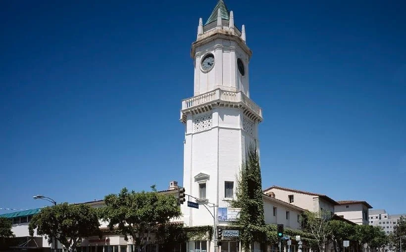A tall white clock tower on a street corner, with trees and buildings nearby, under a clear blue sky.