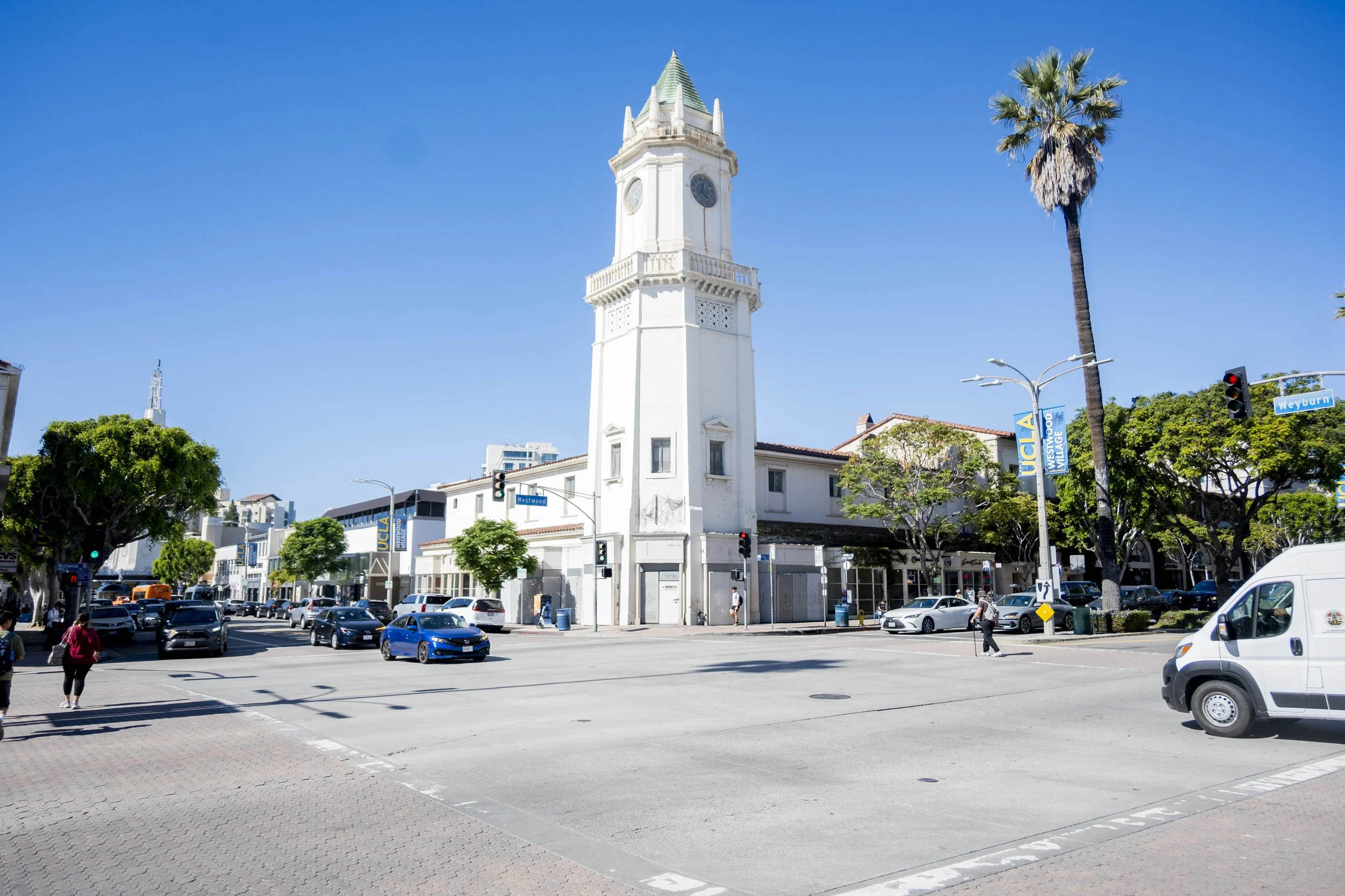 An urban intersection on a sunny day with a white clock tower, palm trees, cars, and pedestrians.