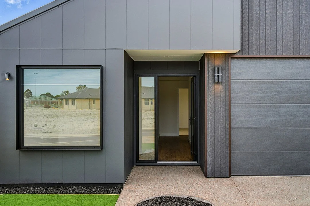 Modern house entrance with black-framed aluminium glass door and window, gray exterior walls, and minimal landscaping.