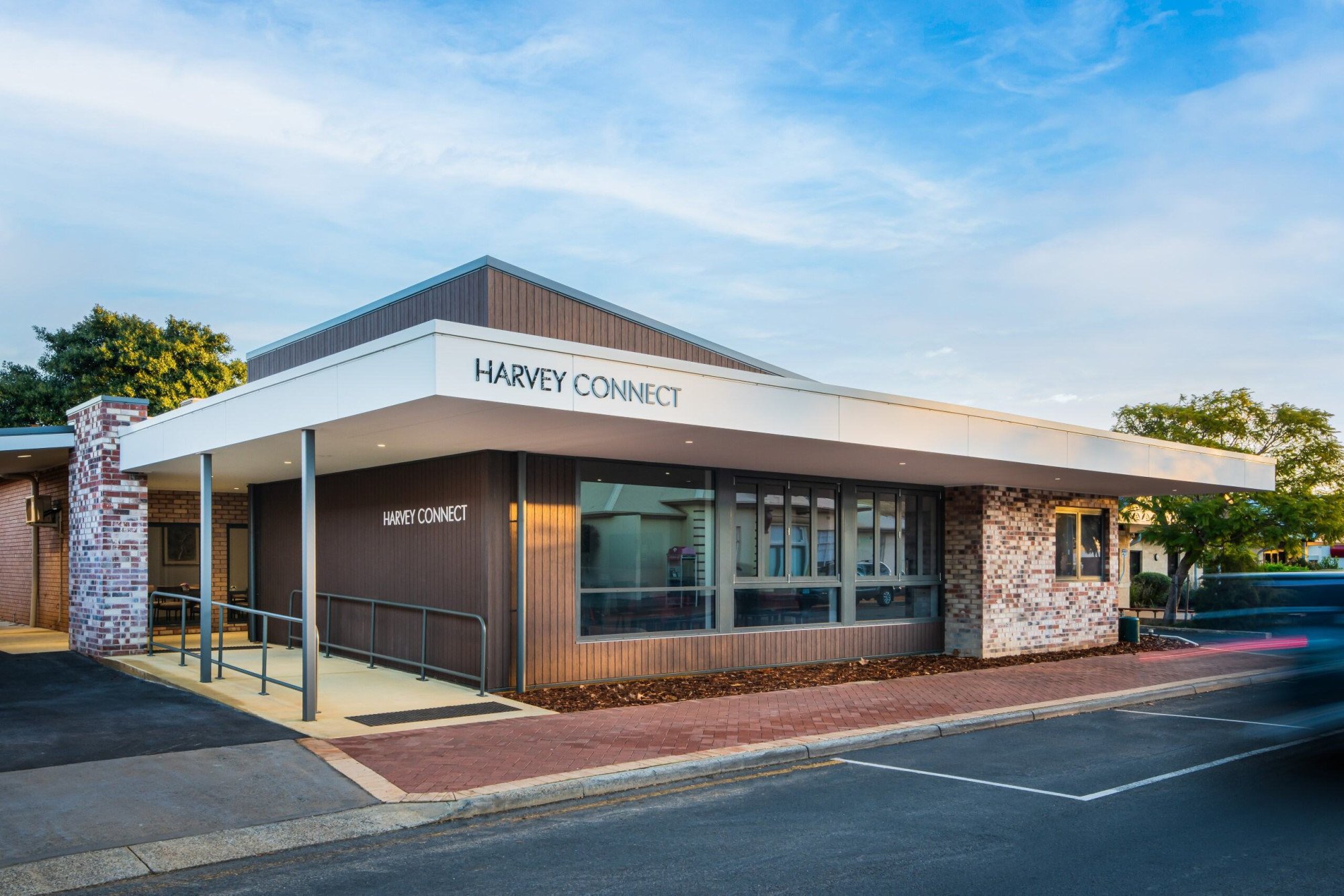 Exterior view of a modern building with the sign 'Harvey Connect' on the white awning and wall, featuring brick and wood paneling, large windows, and a curbside parking lot.