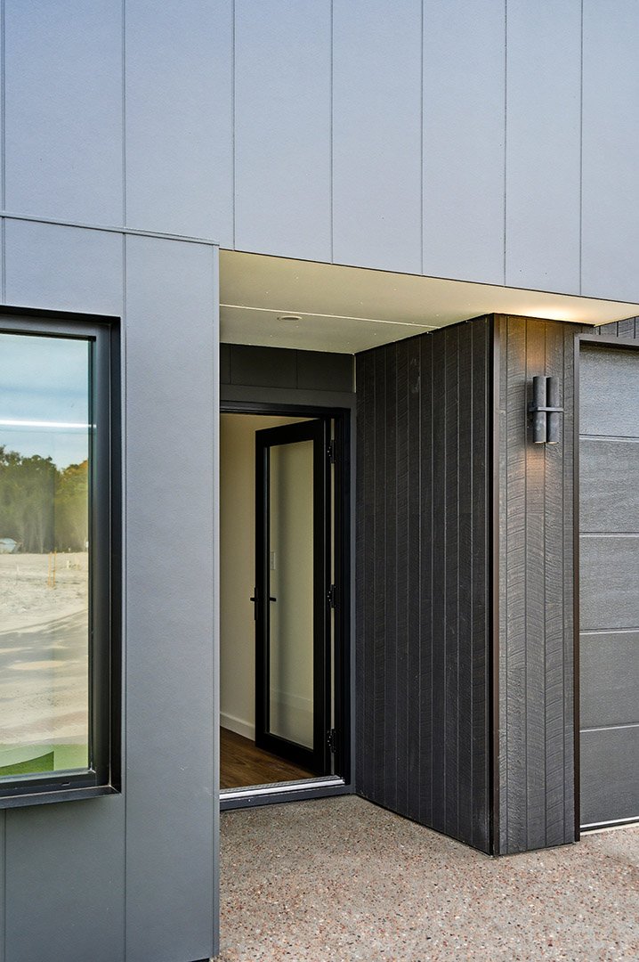 Modern house exterior with gray and black siding, black framed glass door, window, and wall-mounted light fixture.