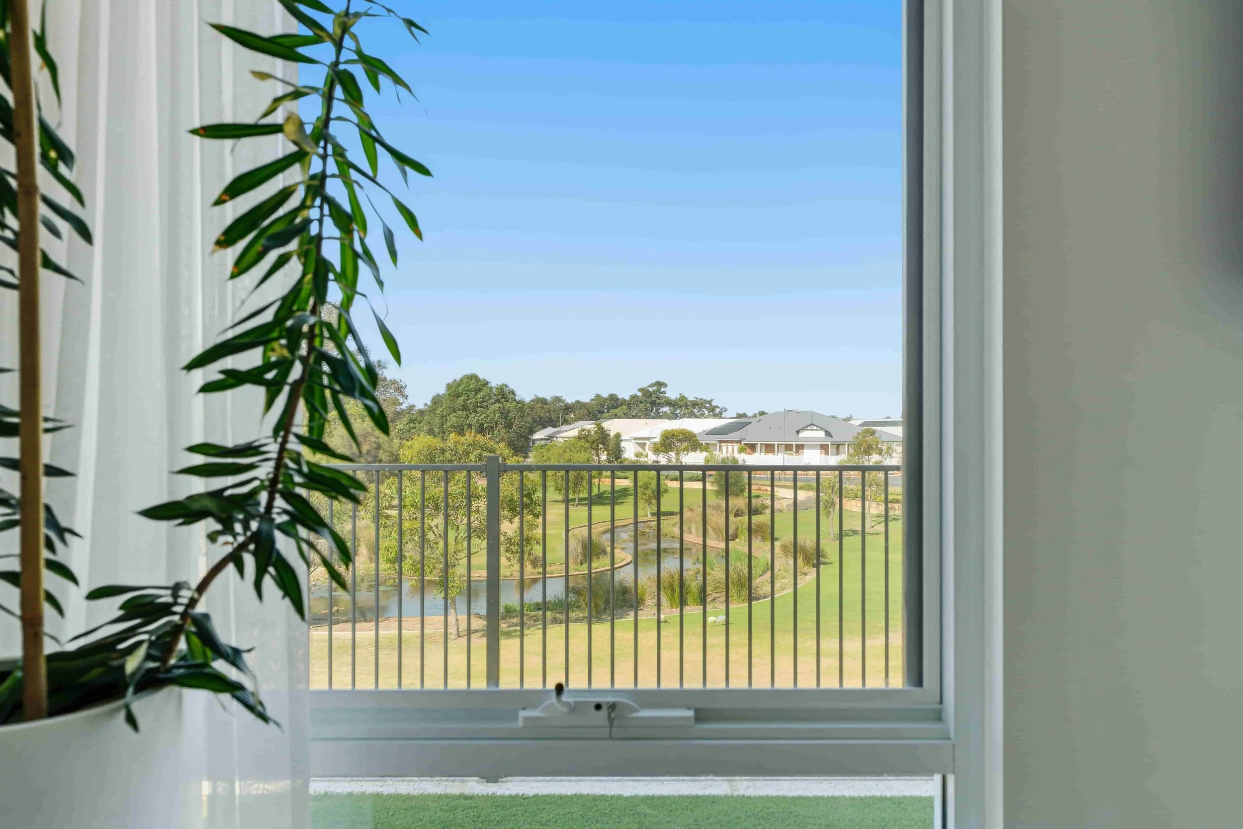 View from inside a room looking out a window at a green landscape with a river, trees, and houses under a clear blue sky.