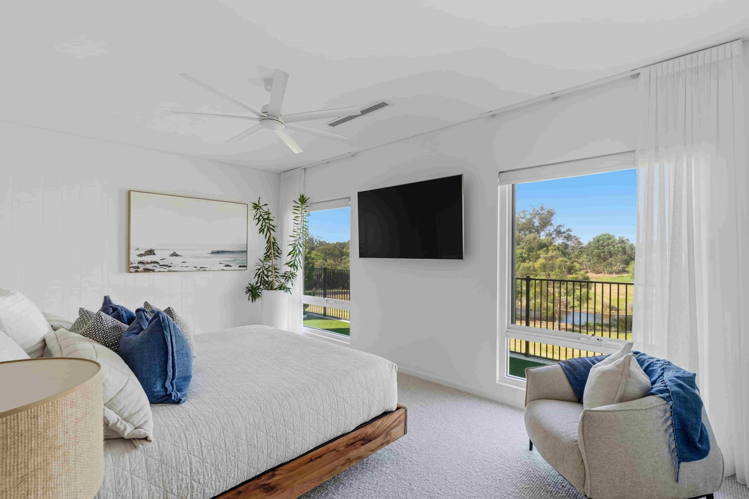 Bright bedroom with white walls, large windows showing outdoor scenery, a bed with white quilt and blue pillows, a beige armchair with a blue throw blanket, a potted plant, wall art, and a television mounted on the wall.