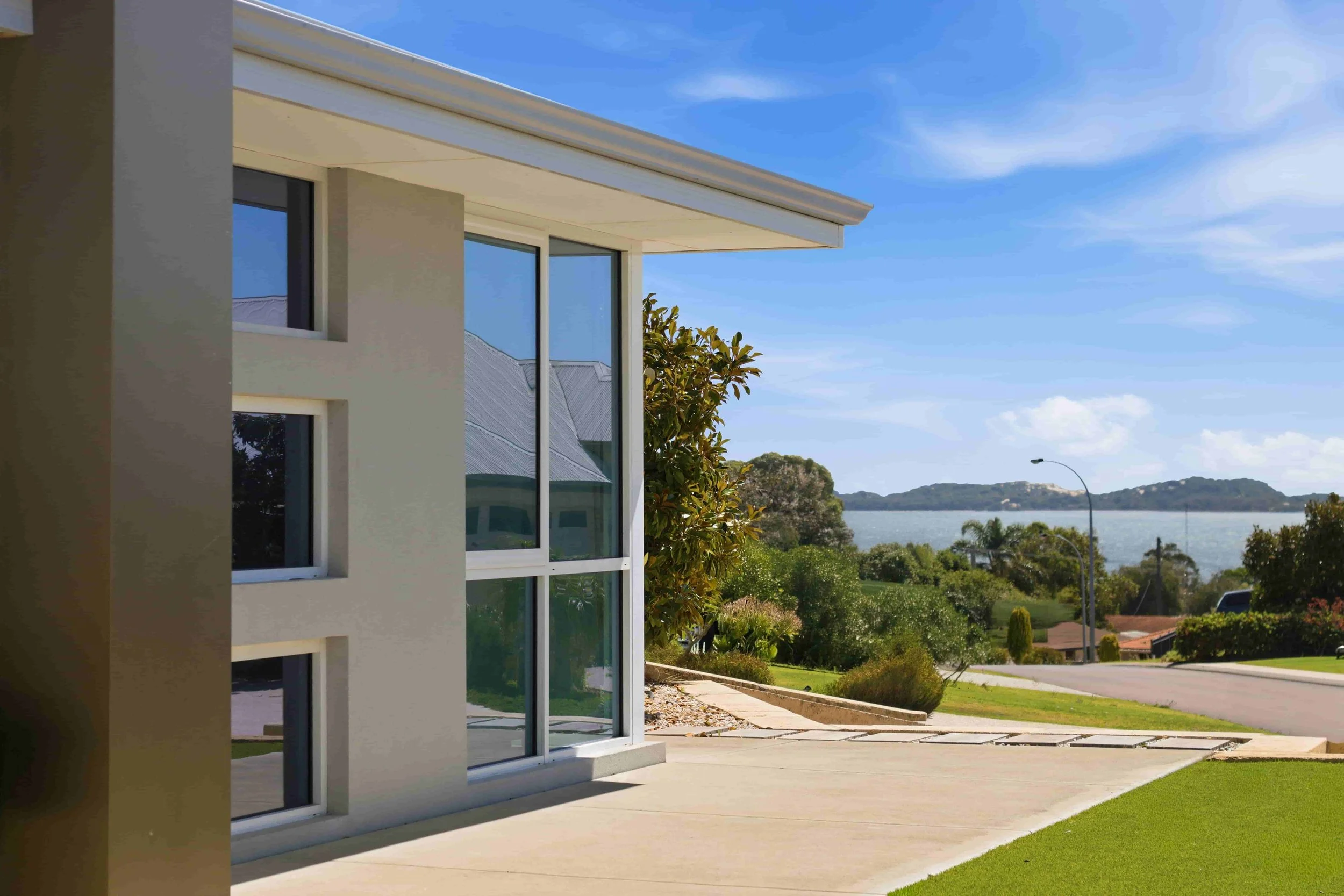 Modern house with large glass windows, front yard, street, greenery, trees, water body, hills, and clear sky.