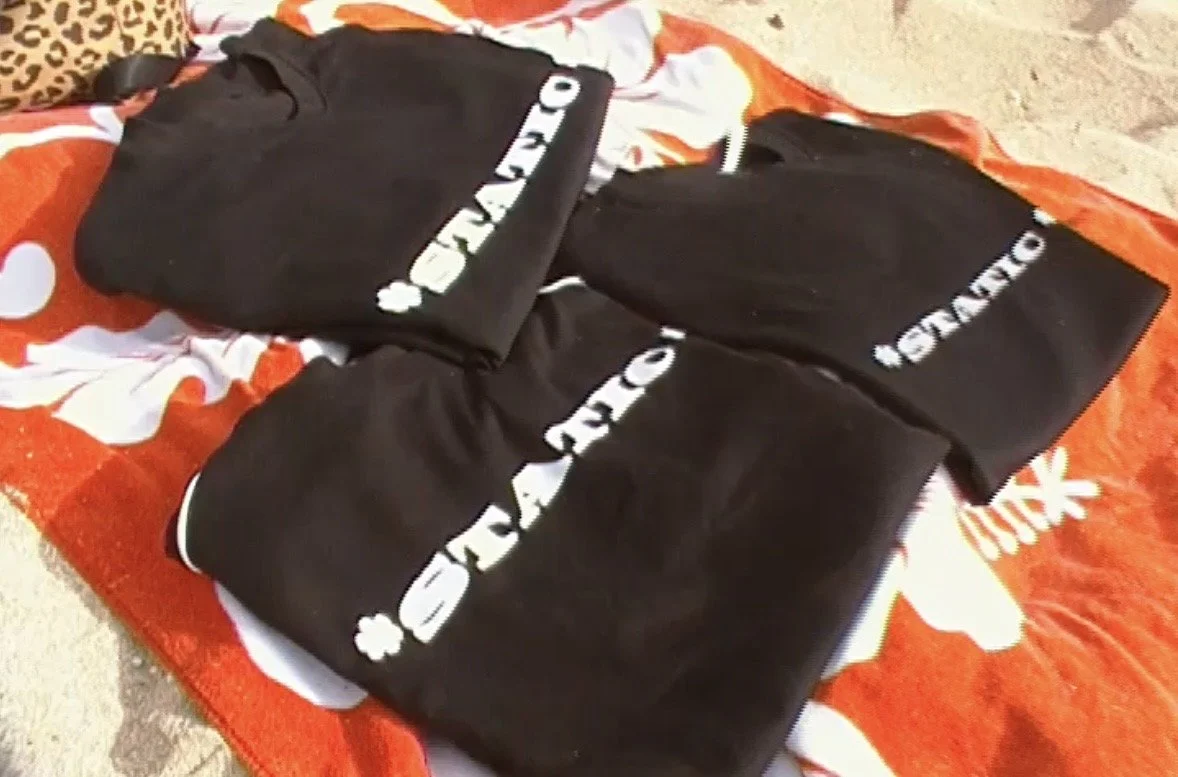 Three black sweatshirts with white text piled on an orange and white towel on the sand.