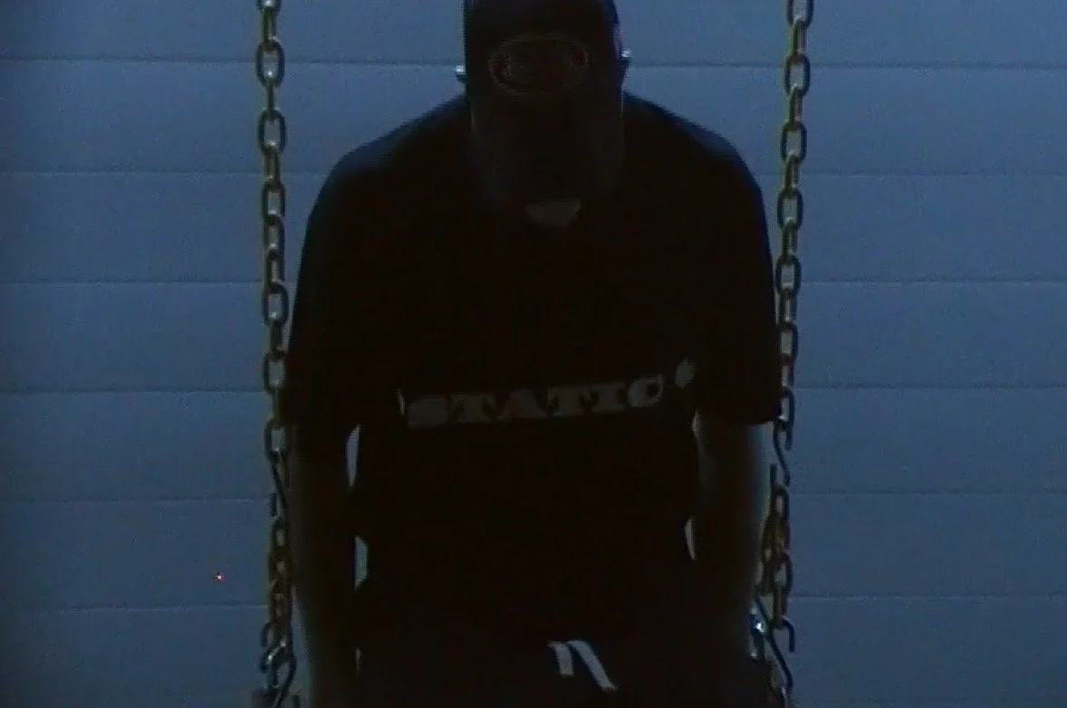 A person sitting on a swing against a blue wall, wearing a dark shirt and a cap, with shadows obscuring their face.