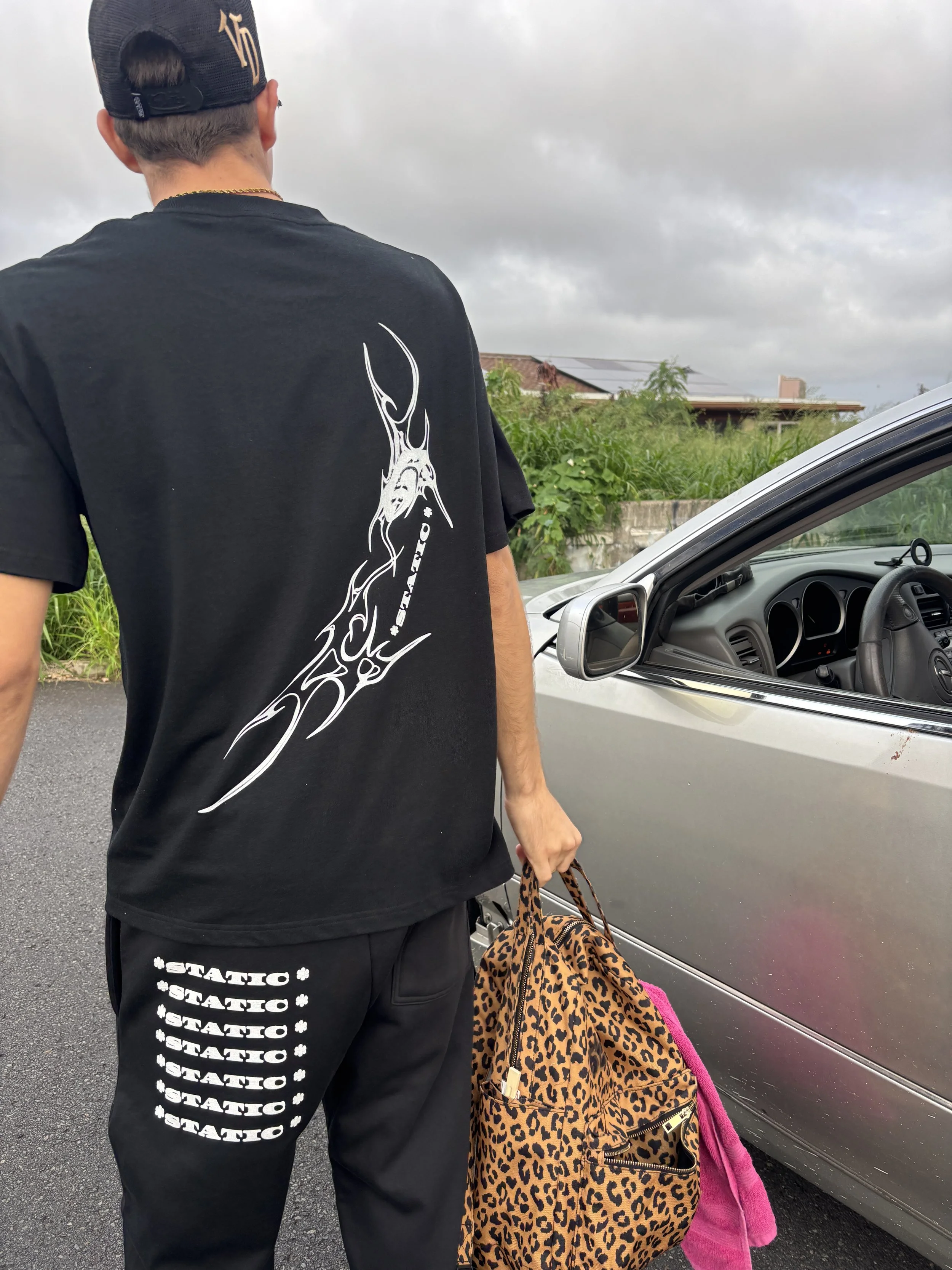 Person standing outdoors near a silver car, holding a leopard print bag and a pink cloth, dressed in a black T-shirt with white graphic design and black pants with 'STATIC' written repeatedly, wearing a black cap.
