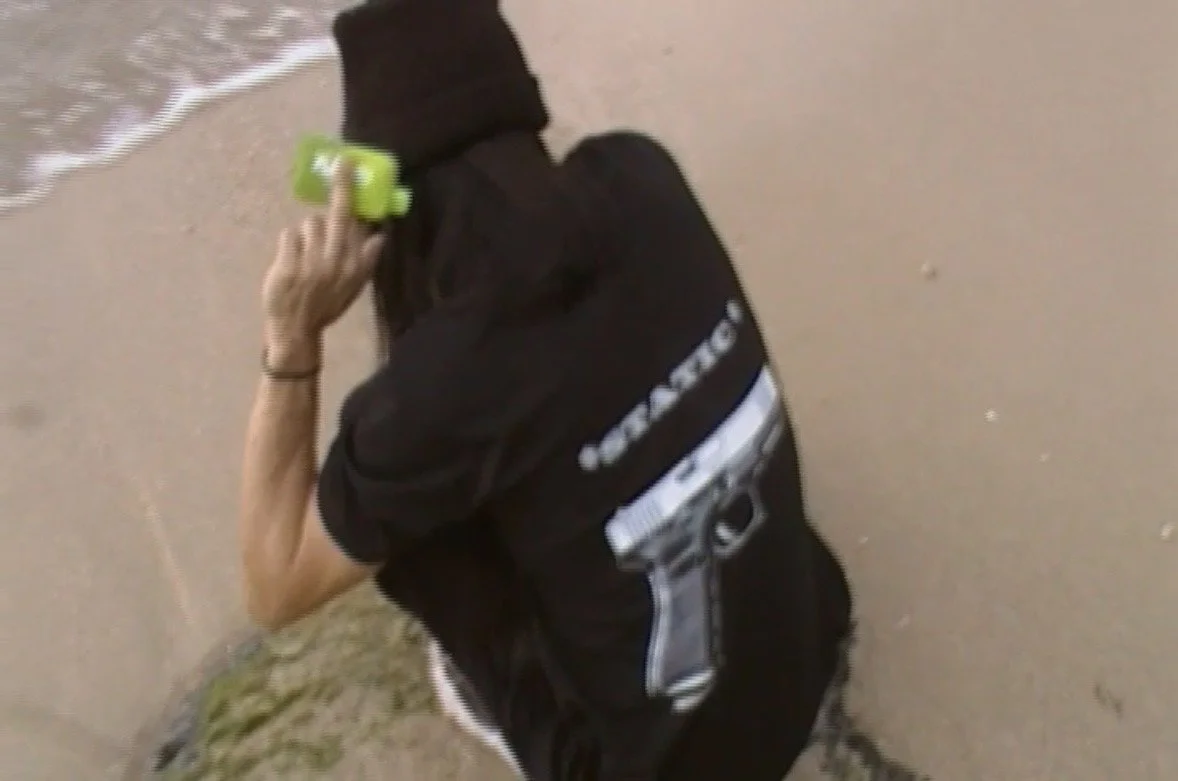 Person in black hoodie kneeling on the sand at the beach, holding a green object to their ear, with the ocean in the background.